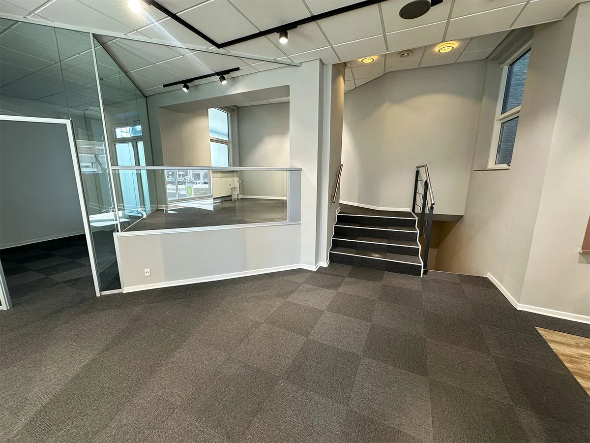 Store lokale med glasdør og trapper til øverste etage. Hvid og grå farvelægning. Moderne indretning med krydsformede loftslys og gulvbelægning.