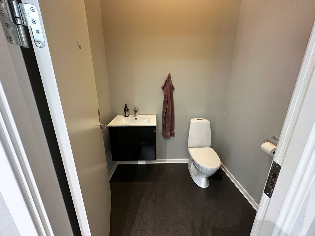 Lejlighed med lille badeværelse og hvid toilet, hvor en håndklæde hænger på væggen. Der er et håndvask med flaske og sæbe. Gulvet er sort og væggene er hvide