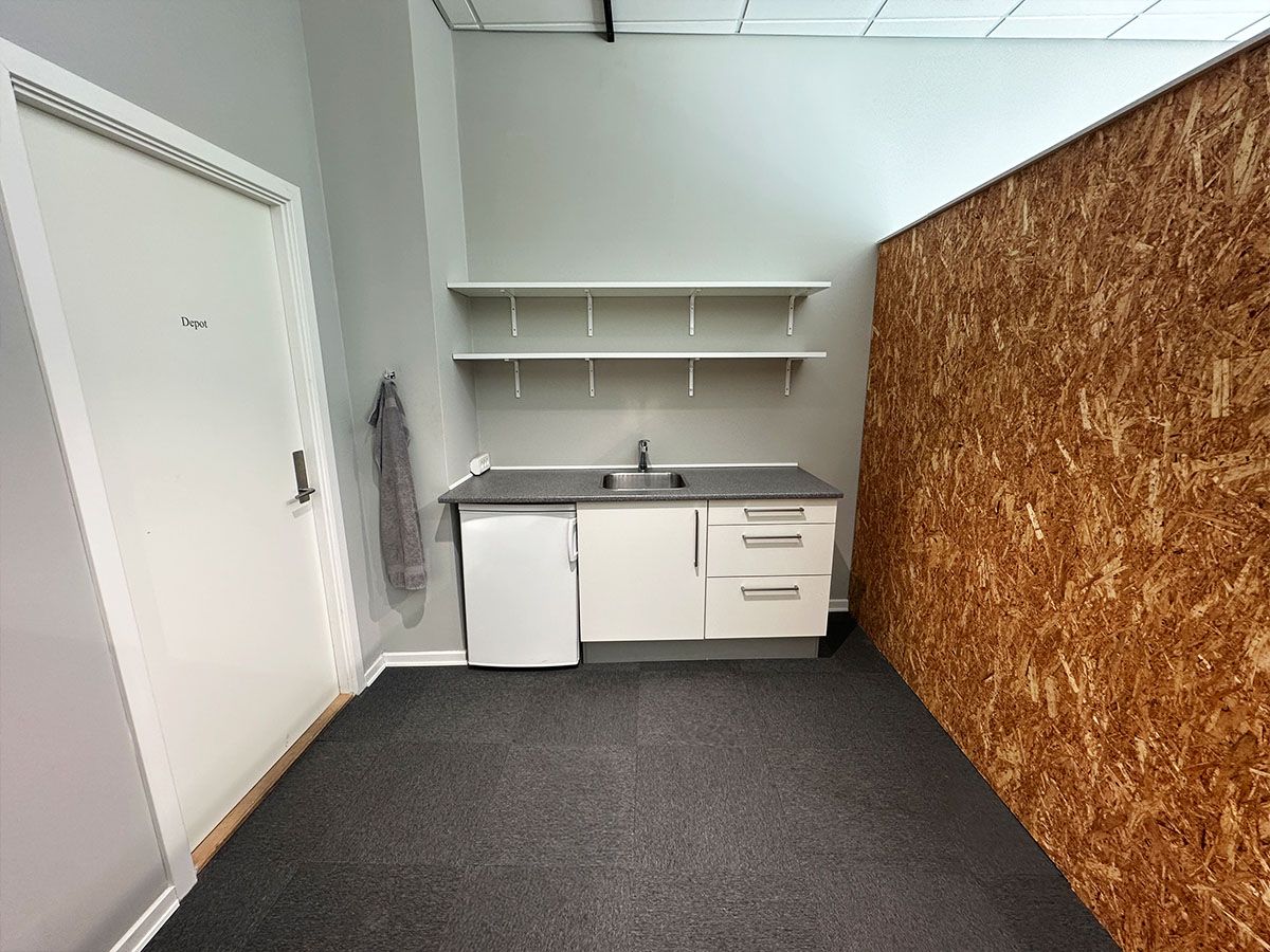 En lille depotrum med hvide vægge og loft, et køkken med lyse, funktionelle hætteskabe, en hvid køleskab og et blikfangsvæg af træplader