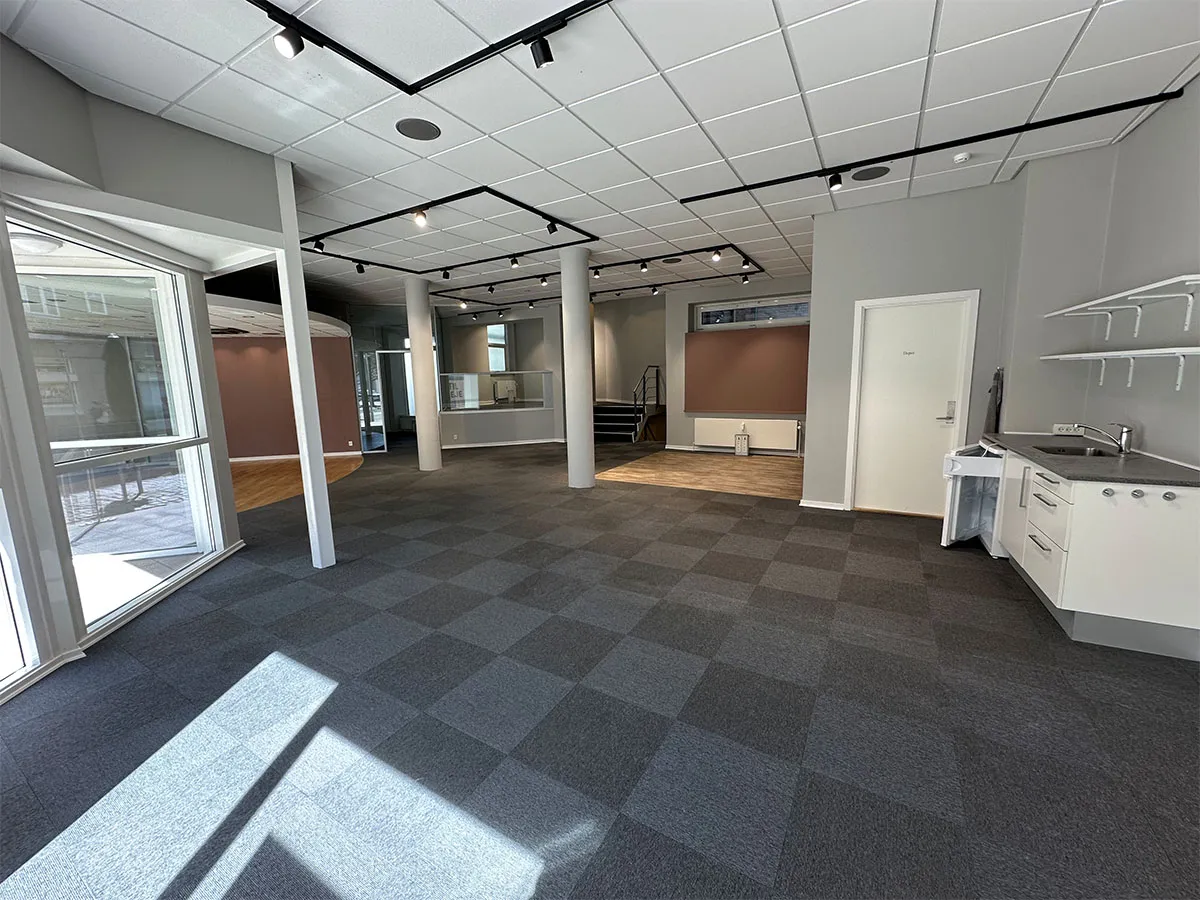 En stor og rummelig stue med højloft, hvide vægge og møbler med simpel design, hvor der er et åbent køkken med moderne møbler og lyse gulvbelægning
