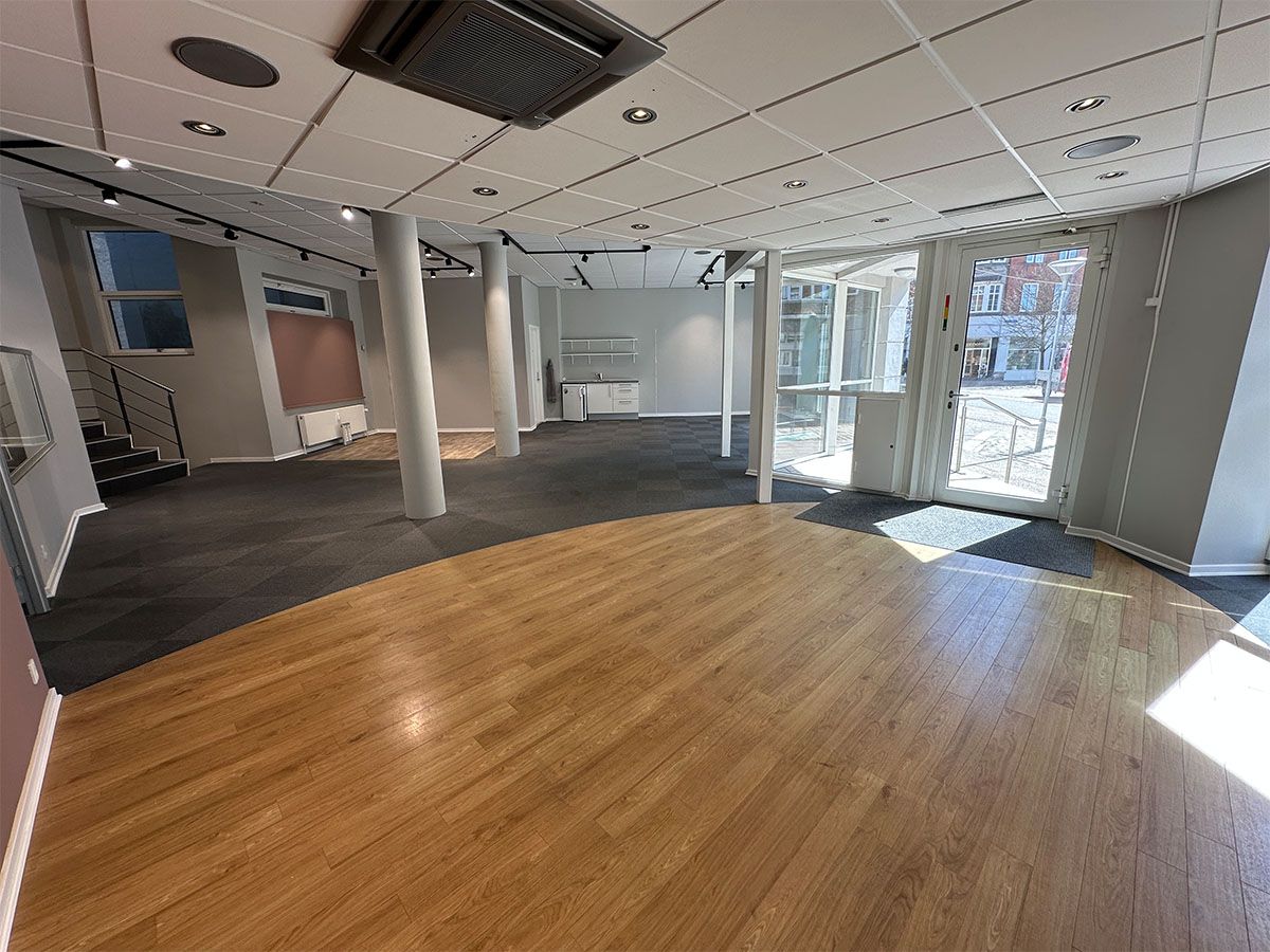 Et stort glasdør- og vinduesanlæg, der fører ud til en gågade. Et kontorlokale med lysegult gulv og hvide vægge. Et sæt trapper med håndtag, som fører ned til første sal.