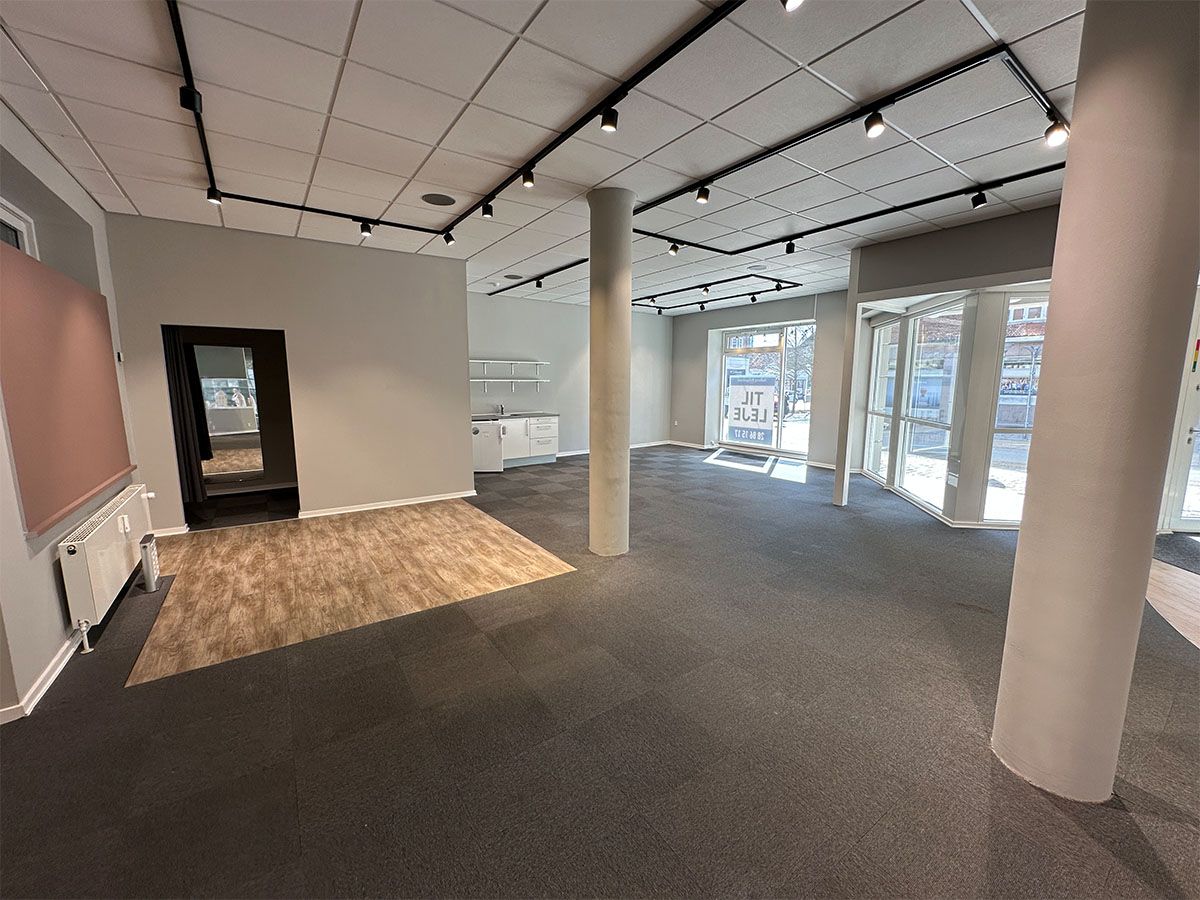 Store lokaler med lyse gulve og hvide vægge. Det er en stor åben stue med flere vinduer og døre. Lamper er monteret i loftet.