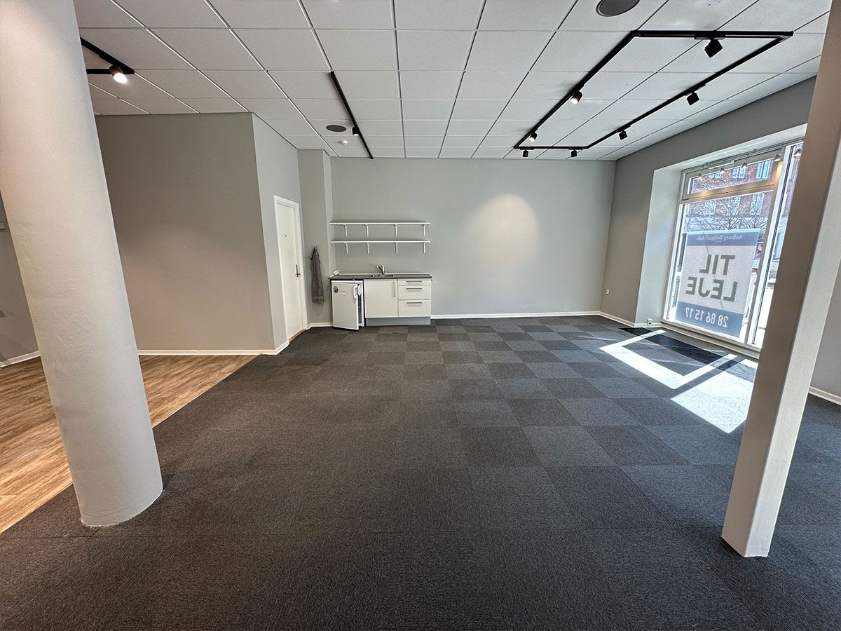 Ledig erhvervslokale med moderne interiør og eksponeret gulvbelægning. Plads til køkken og toilet samt vinduer med reklame for lejlighed. Sorte loftsbøjler med hvide loftsplader.