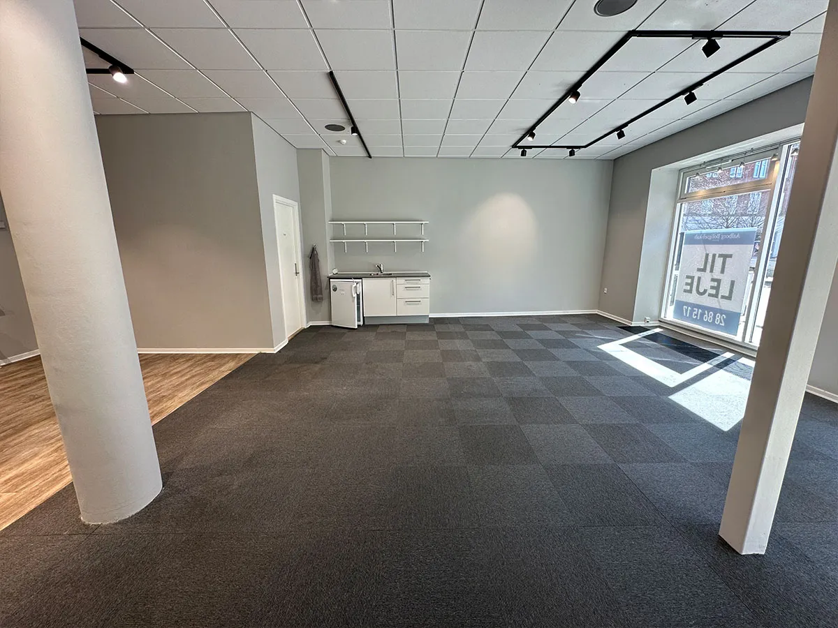 Ledig erhvervslokale med moderne interiør og eksponeret gulvbelægning. Plads til køkken og toilet samt vinduer med reklame for lejlighed. Sorte loftsbøjler med hvide loftsplader.