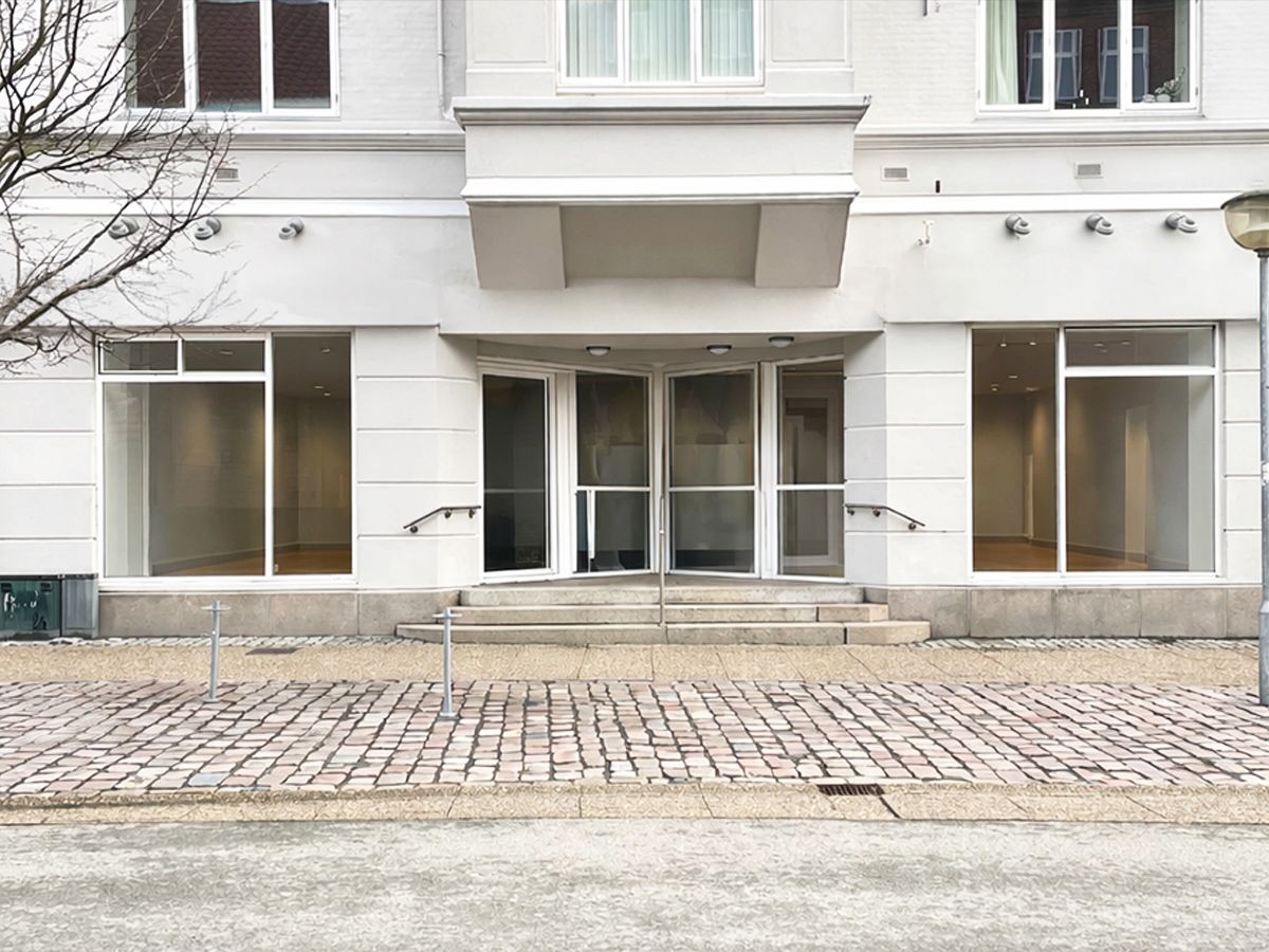 Hjørring bymidte med gågade Østergade, hvor der er en butik med glasvinduer og en hvid facade. Adgangen til butikken er via tre trappetrin og to håndtag