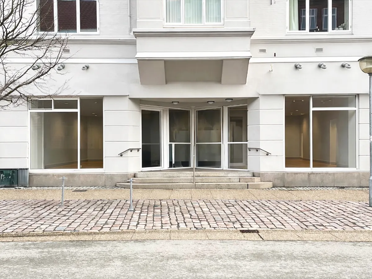 Hjørring bymidte med gågade Østergade, hvor der er en butik med glasvinduer og en hvid facade. Adgangen til butikken er via tre trappetrin og to håndtag