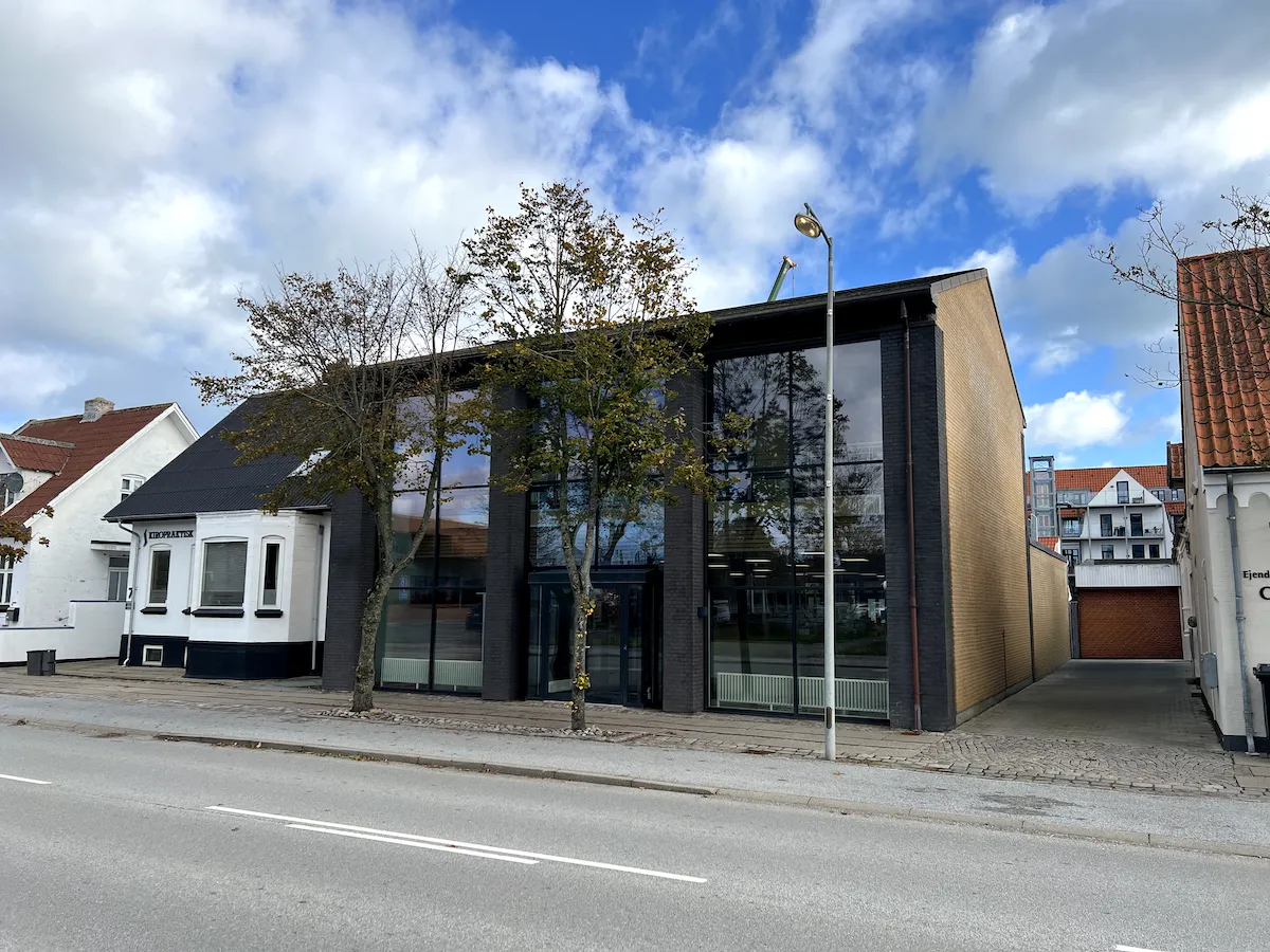 Bygning med store glasruder og sort tag er beliggende på Parallelvej 9 i Frederikshavn. Solrigt vejrum med to træer og gadelampe. Foran er en skrapelejlighed med tegltag og hvide vægge