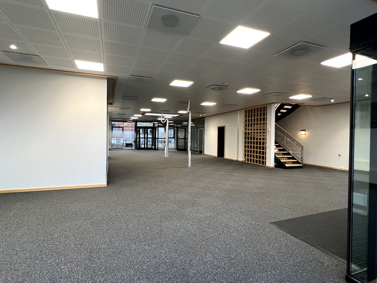 Store, åbne kontor- og butikslokaler med højt loft og indbyggede lyskakter. Der er hvide vægge og et gulvtæppe foran indgangen. Stiger er til højre.