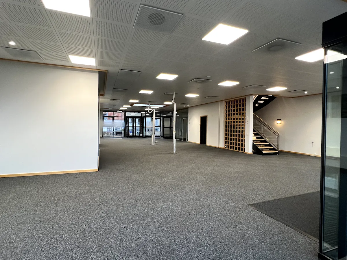 Store, åbne kontor- og butikslokaler med højt loft og indbyggede lyskakter. Der er hvide vægge og et gulvtæppe foran indgangen. Stiger er til højre.