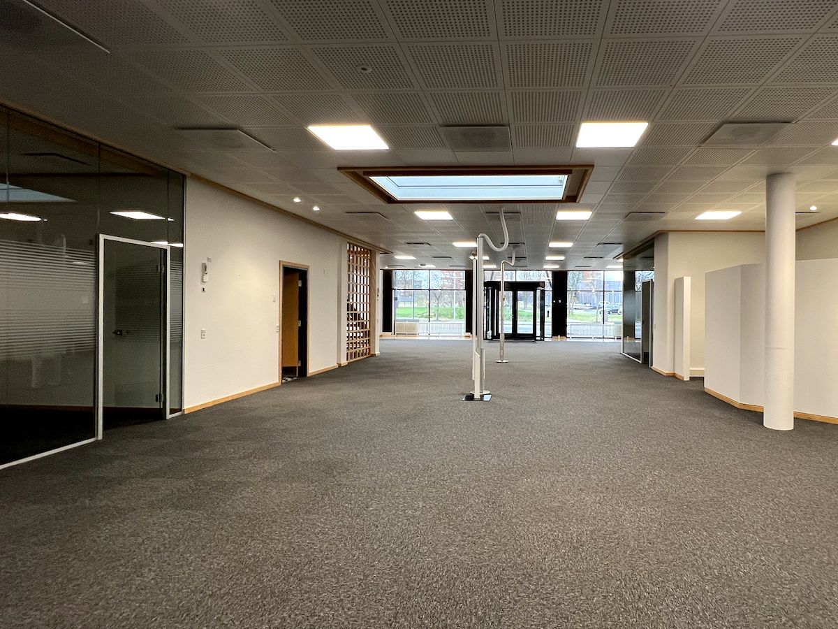 Et stort, lyst og åbent kontorlokale med glasvinduer og døre, hvor der er møbler og indretning. Der er hvide søjler, smukke loftsbelægninger og store vinduer, der giver masser af lys.