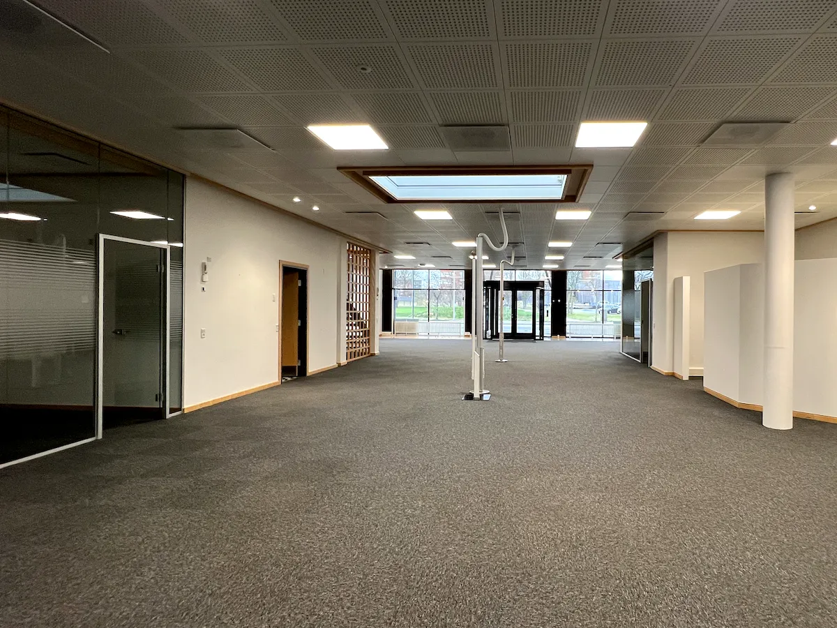 Et stort, lyst og åbent kontorlokale med glasvinduer og døre, hvor der er møbler og indretning. Der er hvide søjler, smukke loftsbelægninger og store vinduer, der giver masser af lys.