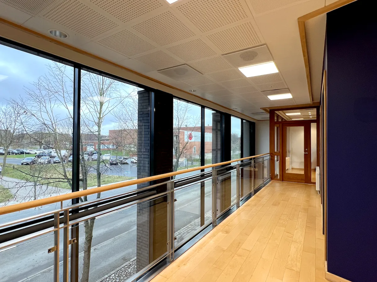 Lejet lokaler med glasparti og trægulv i Frederikshavn med udsigt til parkeringsplads og vej samt lyse loftslamper og møblerede kontorrum