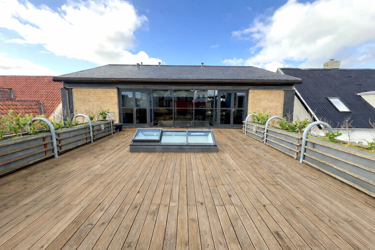 Terrasse med udsigt til Parallelvej 9 i Frederikshavn. Trægulv med skyggefulde planter og glasdøre til bygning med rødt tag