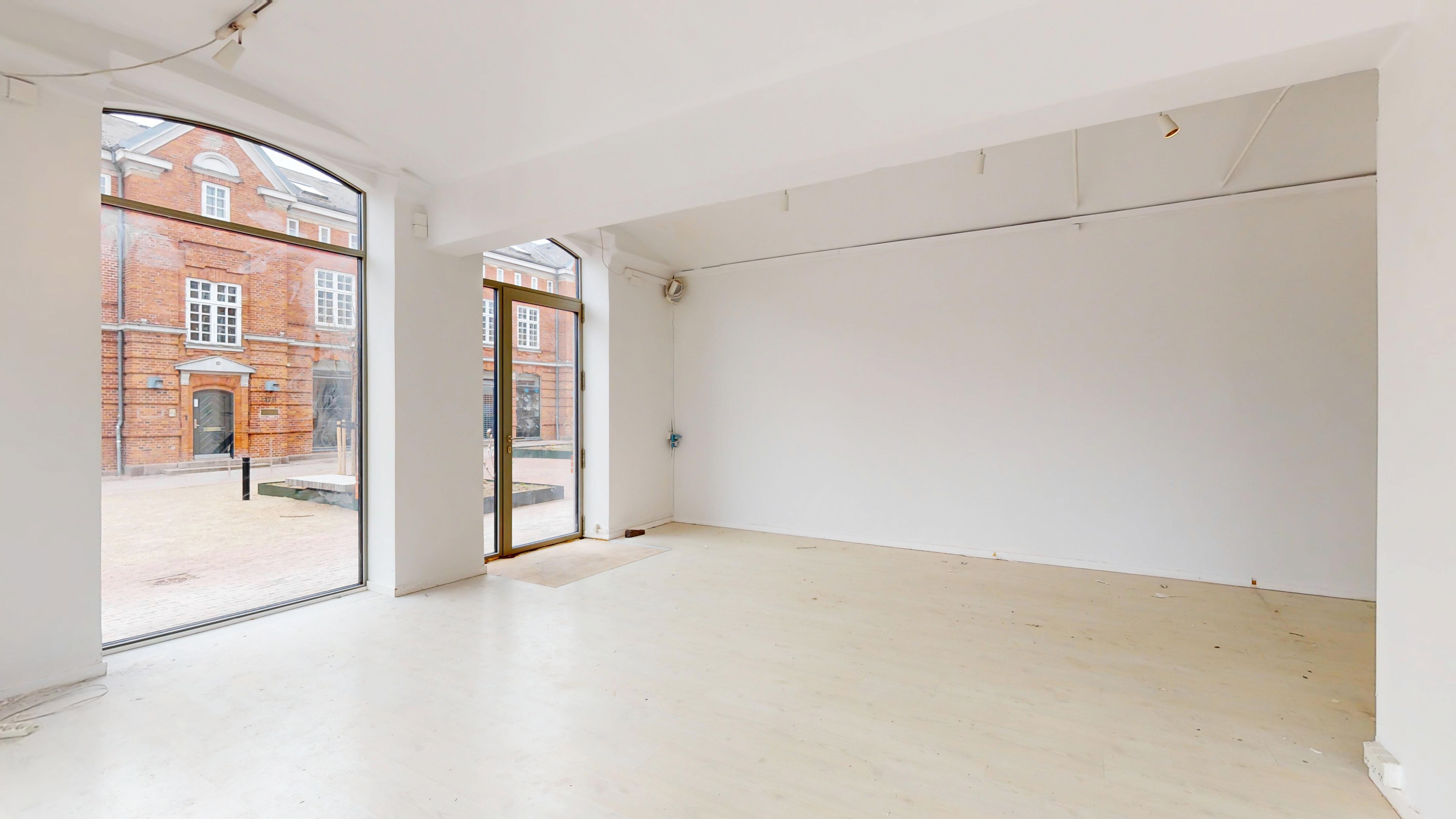 Lejlighed med stort glasvindue og dobbeltdør. Hvidt loft og vægge. Bred gulv. Bygning med røde mursten og vinduer i baggrunden.