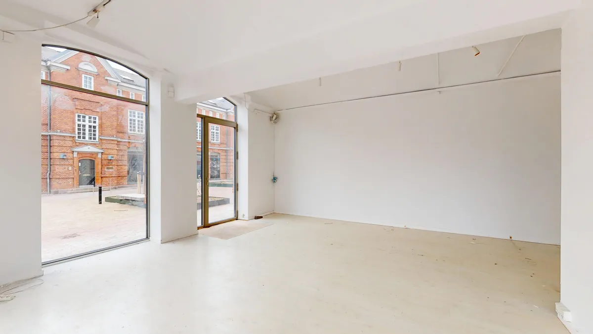 Lejlighed med stort glasvindue og dobbeltdør. Hvidt loft og vægge. Bred gulv. Bygning med røde mursten og vinduer i baggrunden.