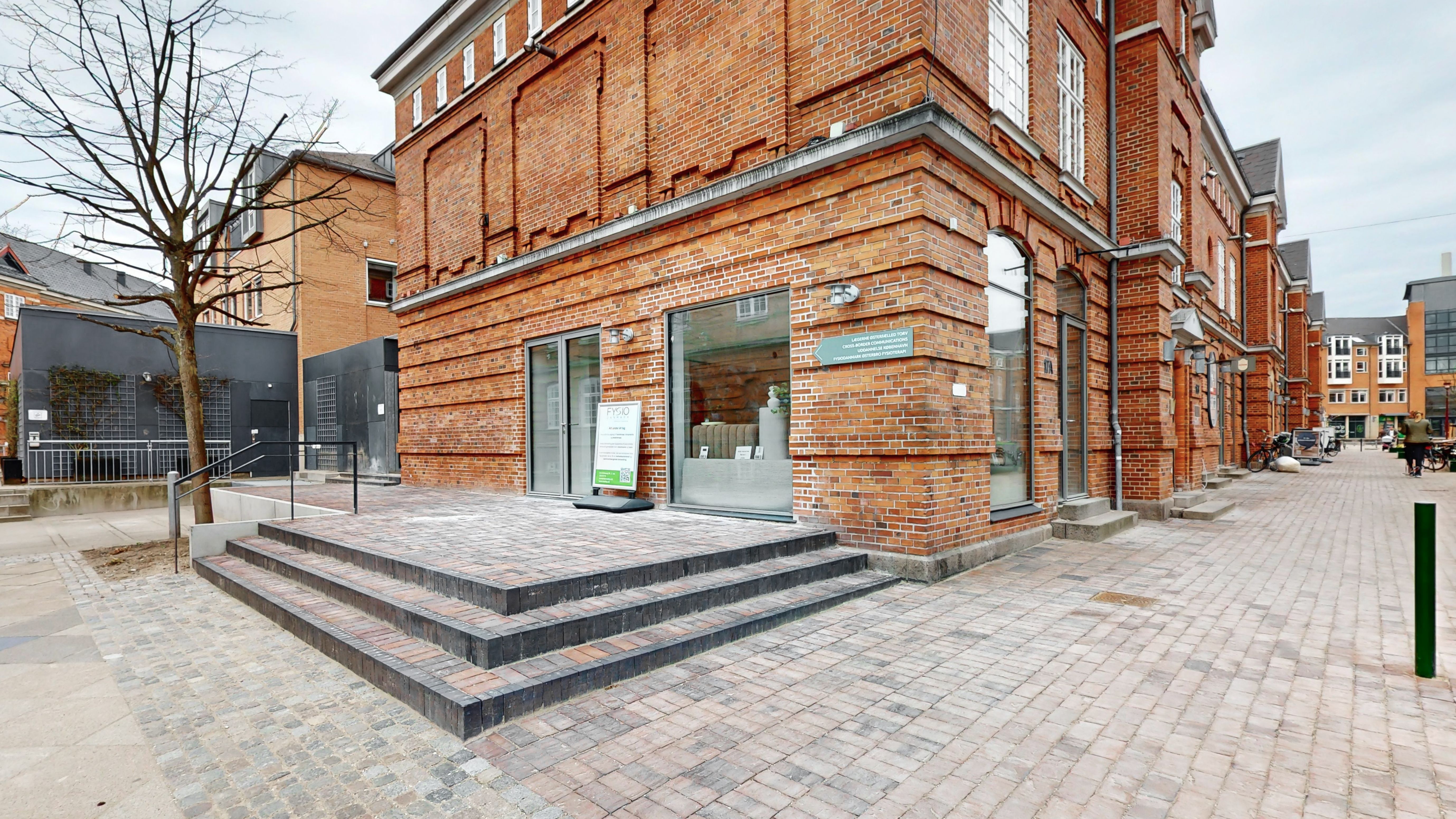 Lejlighed i røde mursten med glasvinduer og store trin udenfor, gård med grønne planter og skulptur, skrappe bygninger og gågade med folk og cykler