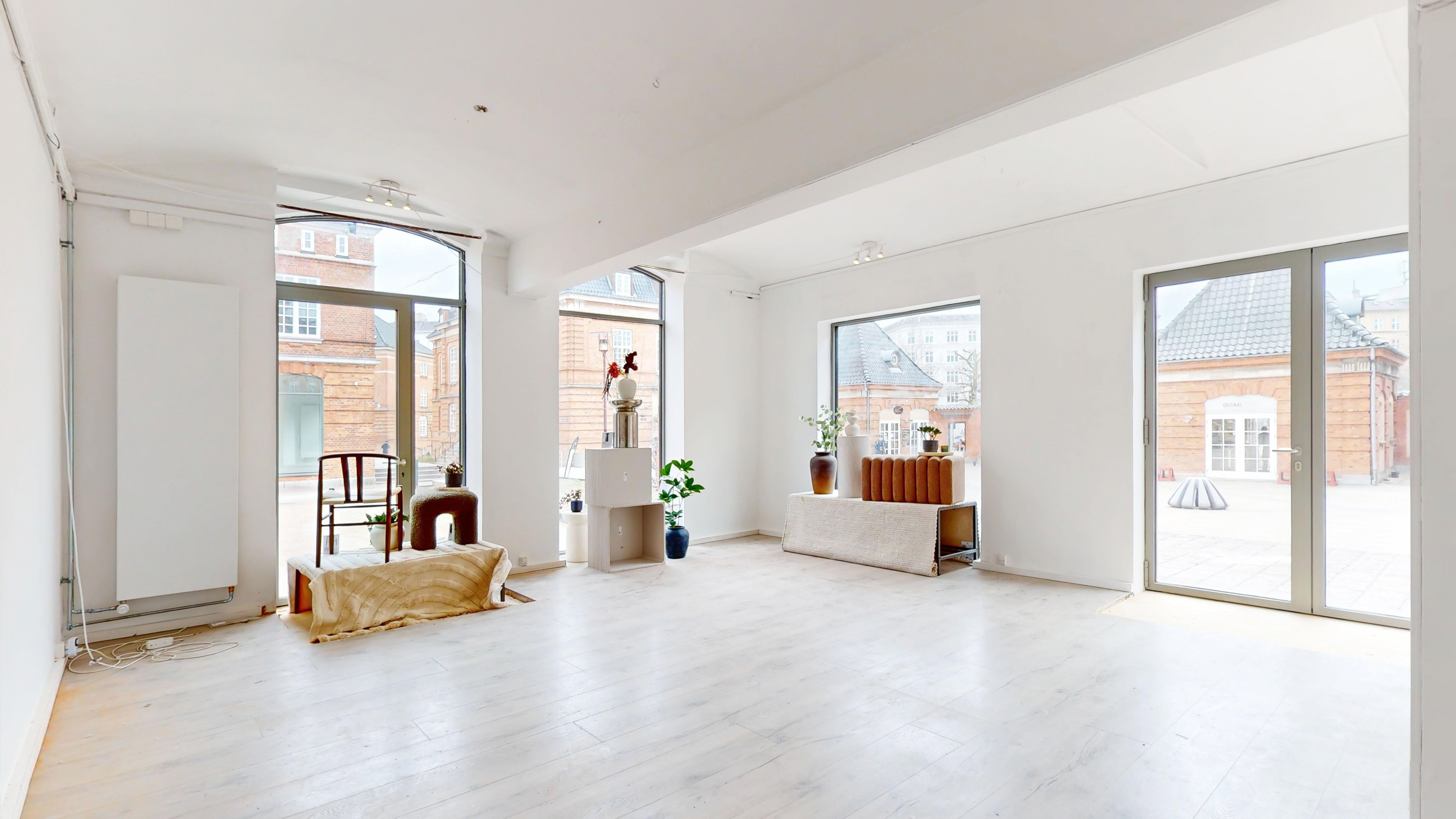 Lyst og indbydende værelse med hvide vægge, trægulv og store vinduer med udsigt til byen og en have. Ingen mennesker er synlige på billedet