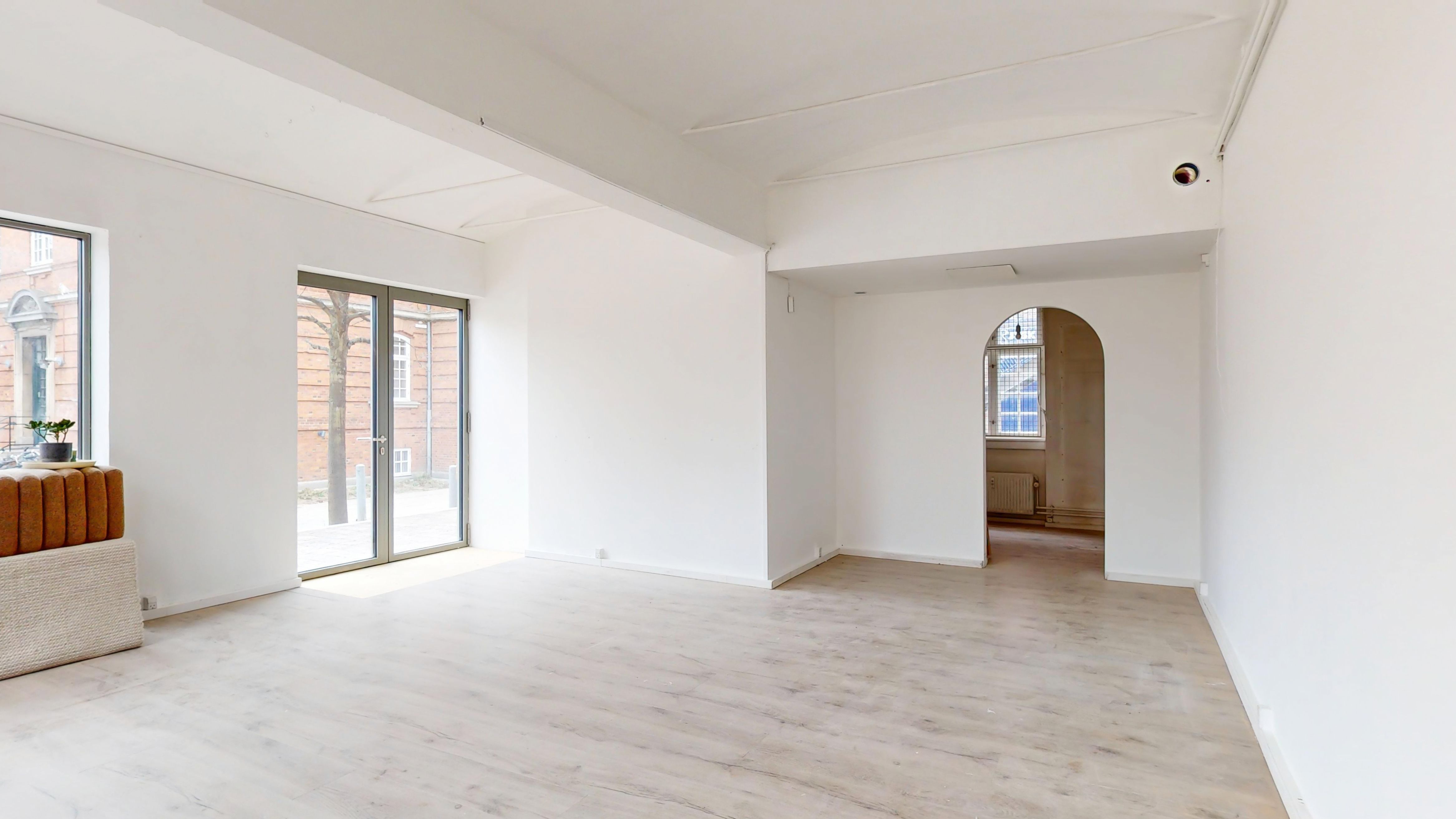 Et lyst og indbydende rum med hvide vægge og lyse gulve, en plante og en åbning mod et lyskapel med ruder