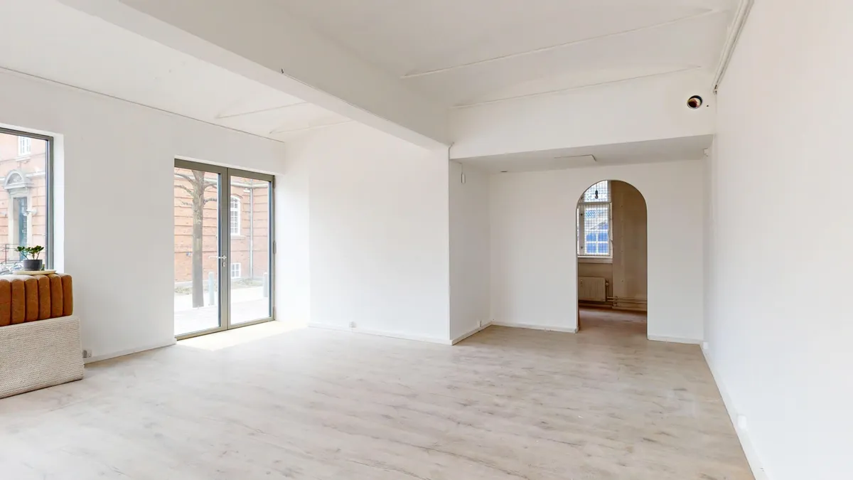 Et lyst og indbydende rum med hvide vægge og lyse gulve, en plante og en åbning mod et lyskapel med ruder