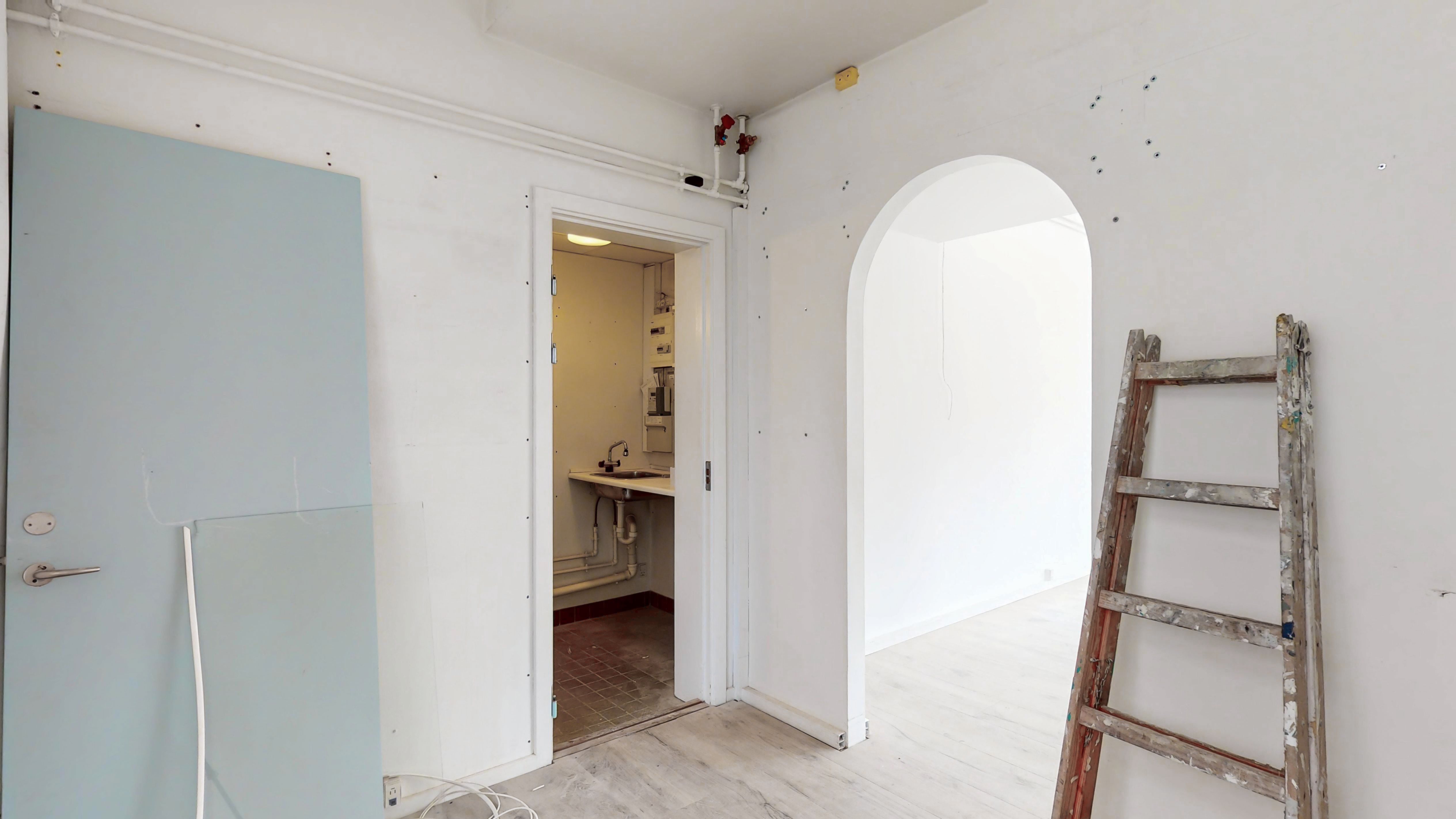 Lyst og rent rum med hvide vægge, trægulv og åben dør til et lille badeværelse med en håndvask og skur. En stige står mod væggen.