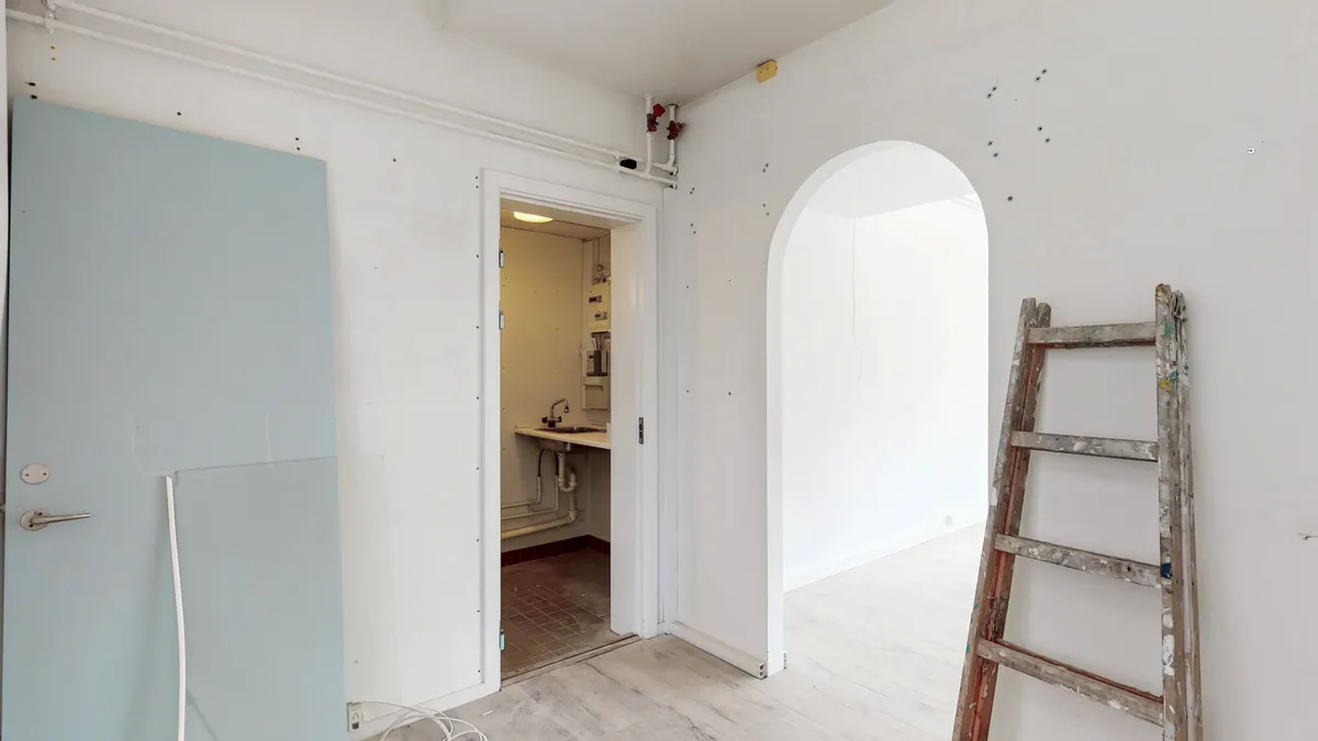 Lyst og rent rum med hvide vægge, trægulv og åben dør til et lille badeværelse med en håndvask og skur. En stige står mod væggen.