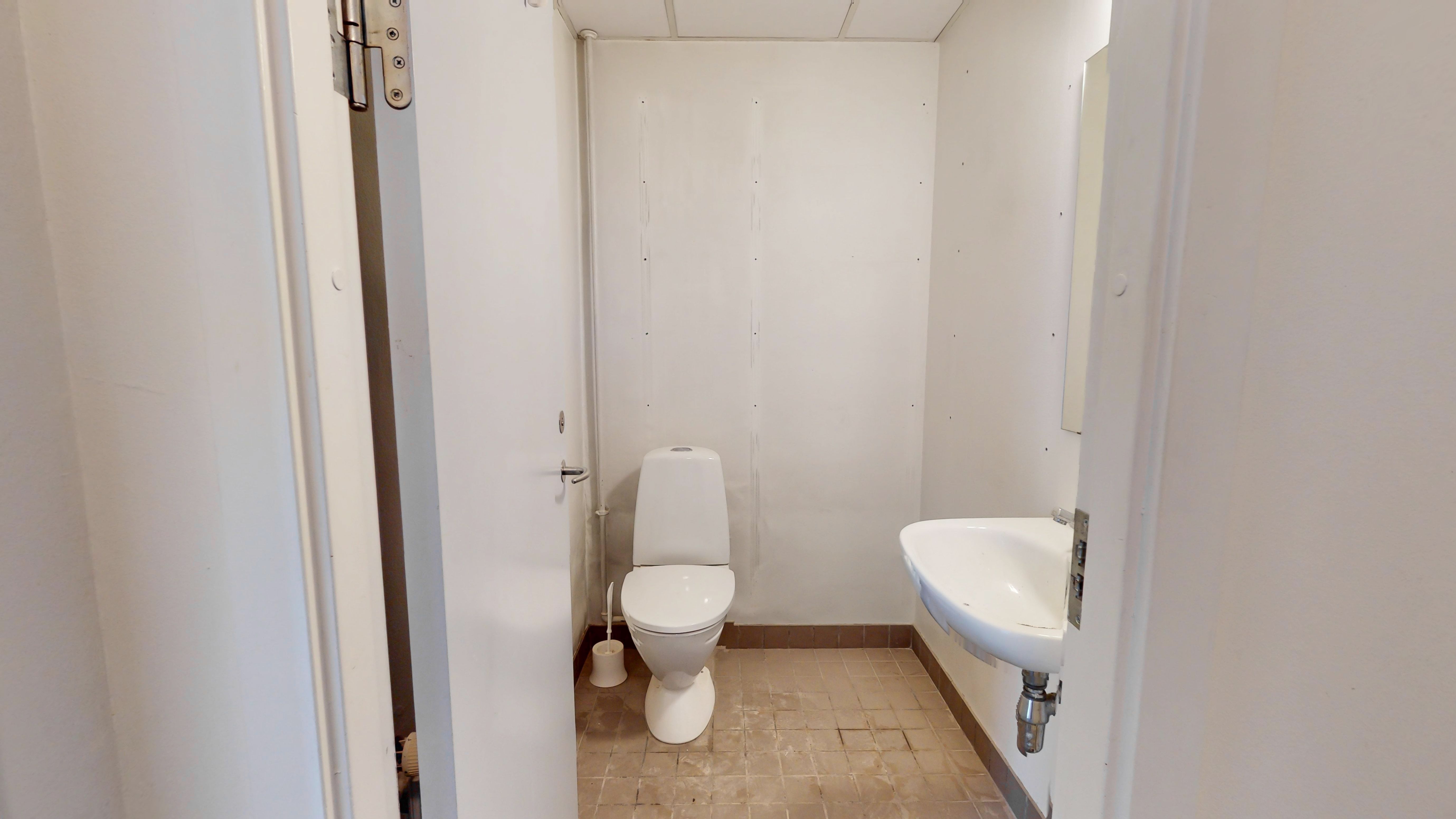 Et lille og funktionelt badeværelse med hvid toilet, hvid vask og spejl samt toiletbørste. Gulvet er dækket med lysebrune fliser.