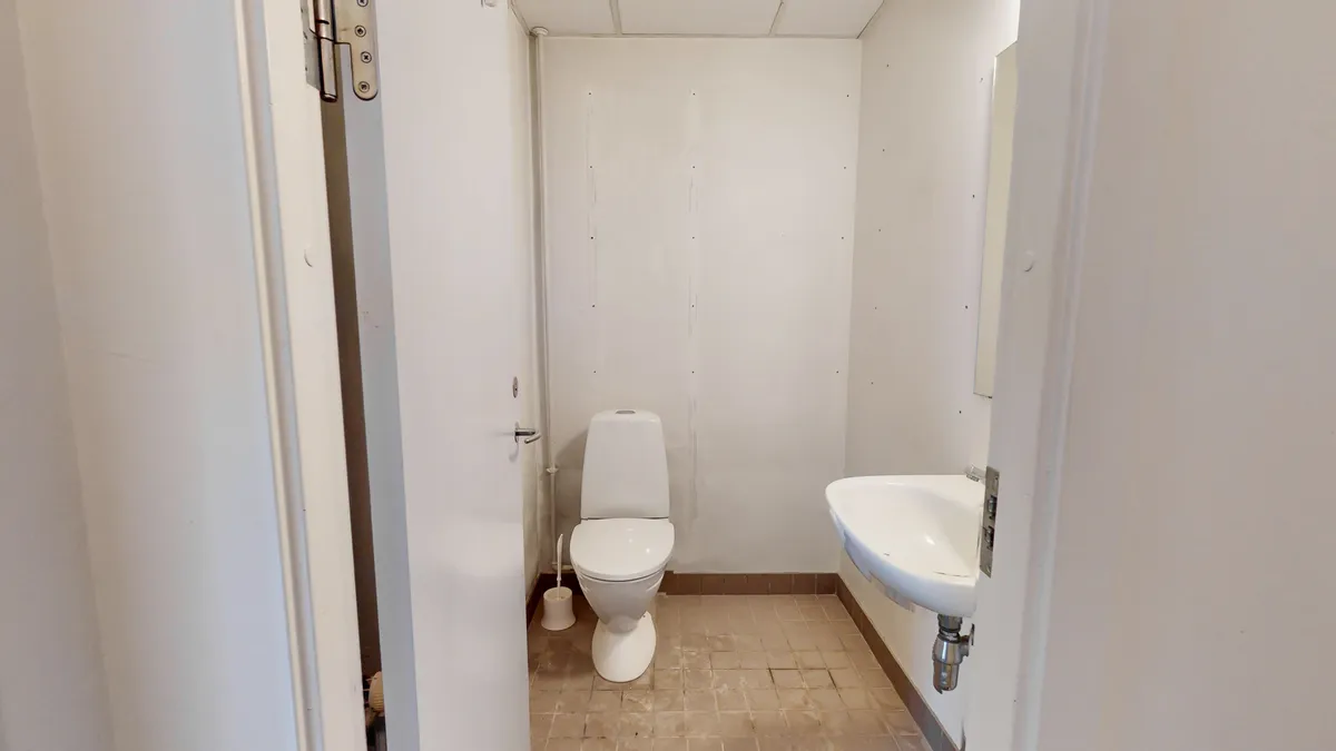 Et lille og funktionelt badeværelse med hvid toilet, hvid vask og spejl samt toiletbørste. Gulvet er dækket med lysebrune fliser.