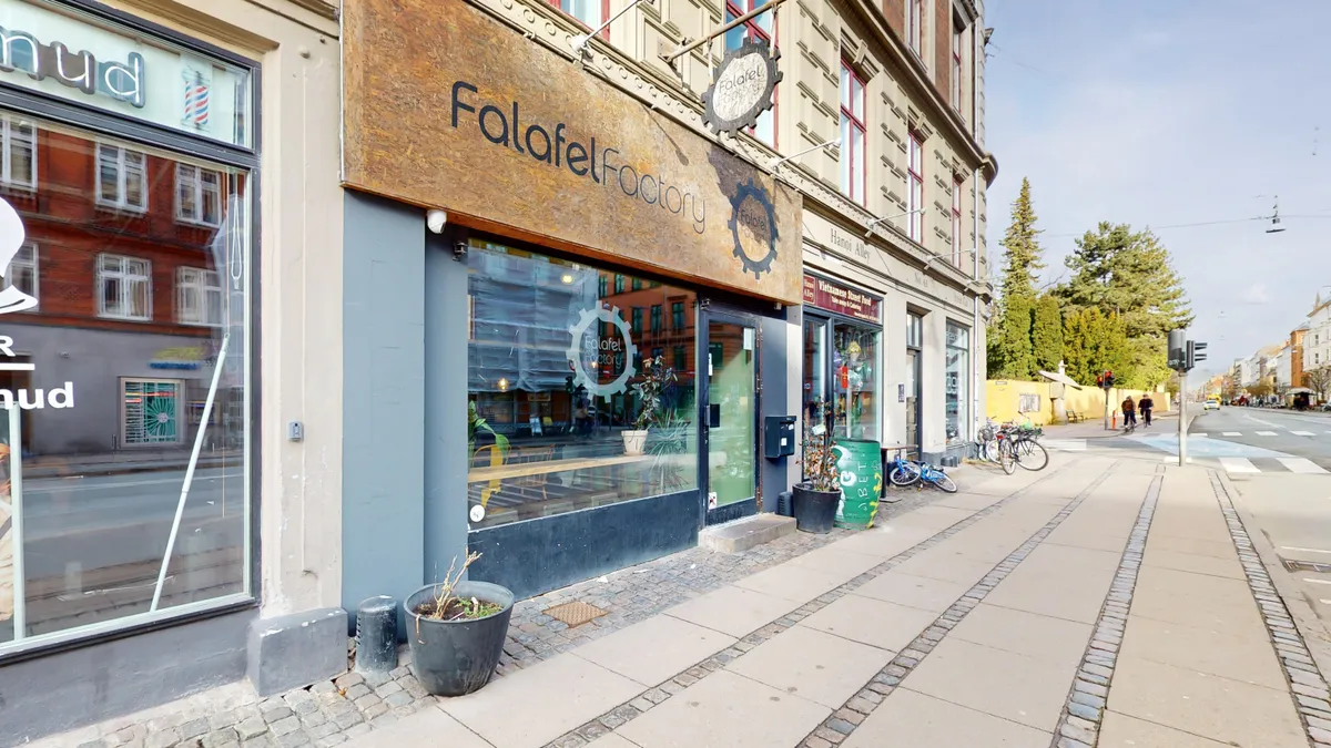 Et moderne falalel-restaurant med facade i træ og metal, med store glasvinduer og udendørs terrasse. En grøn spand og en grøntplante står på fortovet.