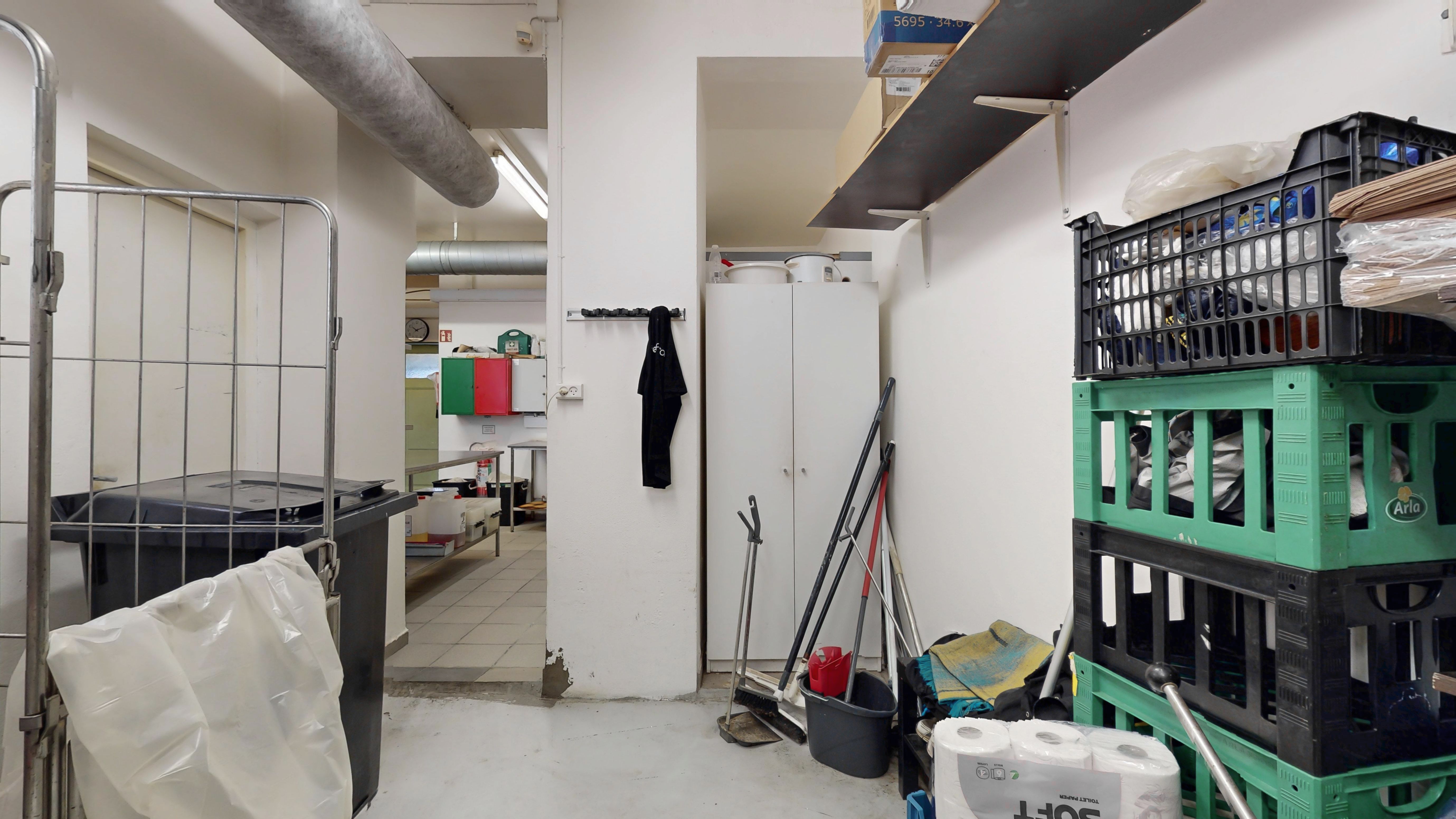 Lagerrum med røde og grønne paller, en hvid køleskab og en stor plastkasse med tøj på toppen. Et skraldespand med plastpose, en svejsermaskine og en røgdetektor er placeret i rummet