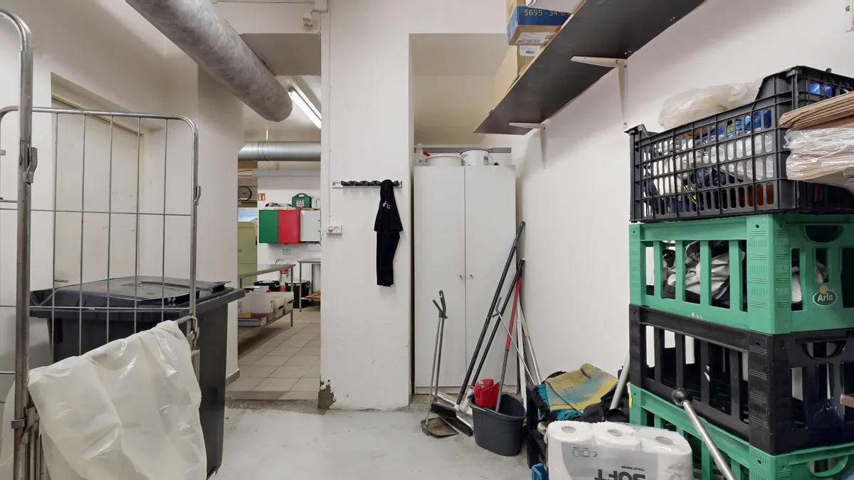 Lagerrum med røde og grønne paller, en hvid køleskab og en stor plastkasse med tøj på toppen. Et skraldespand med plastpose, en svejsermaskine og en røgdetektor er placeret i rummet