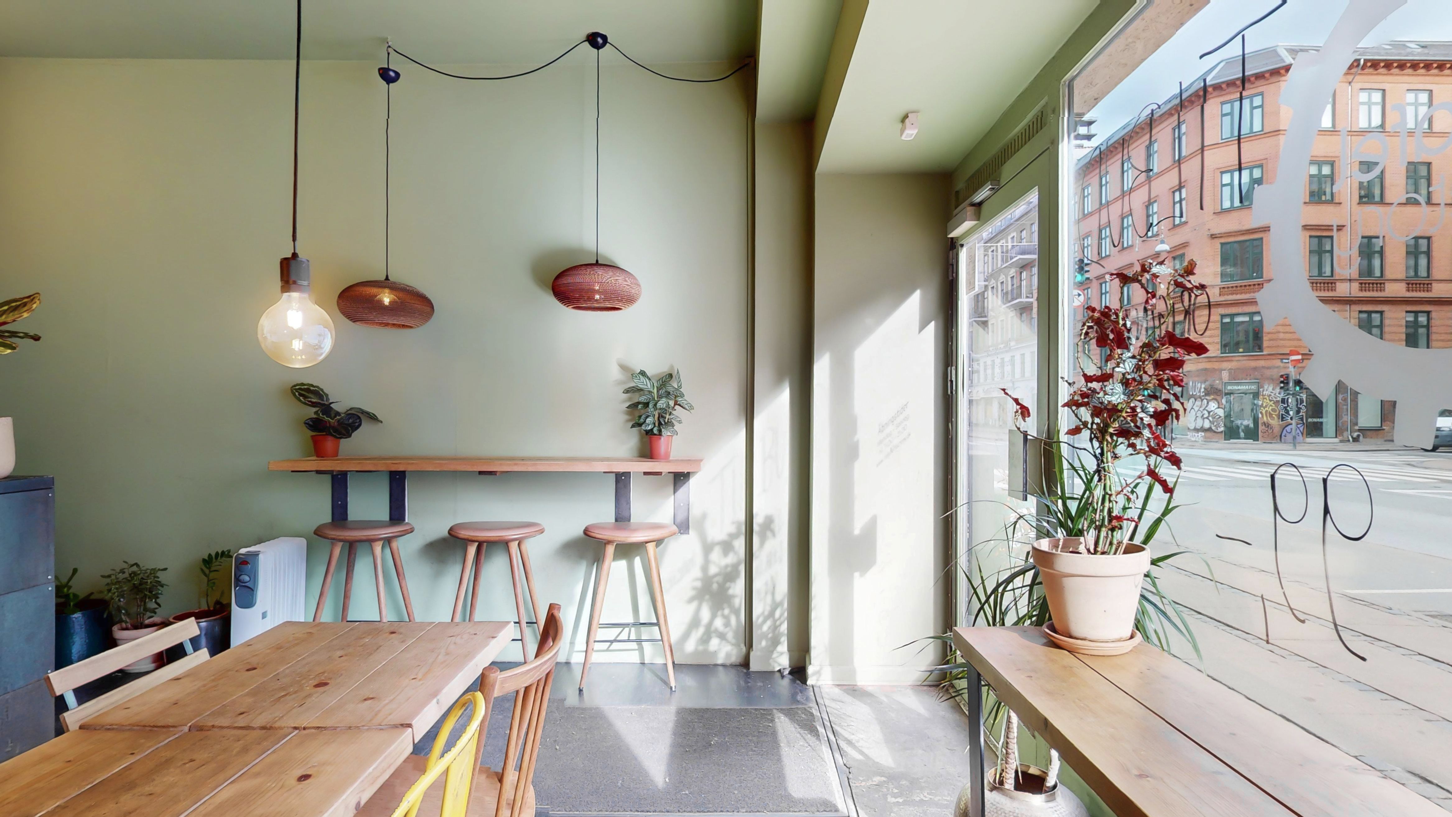 Et lyse interiør med træborde og stole samt plantepoter og lysekroner samt udsigt til gaden med bygninger og trafik