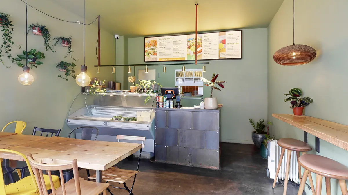 Restaurant med plantegred og lyse loftslamper, salgsskilt med hjemmelavet pizza, bord og stole, salgsdisk med plantegred og flere planter samt to stolhynder med stol