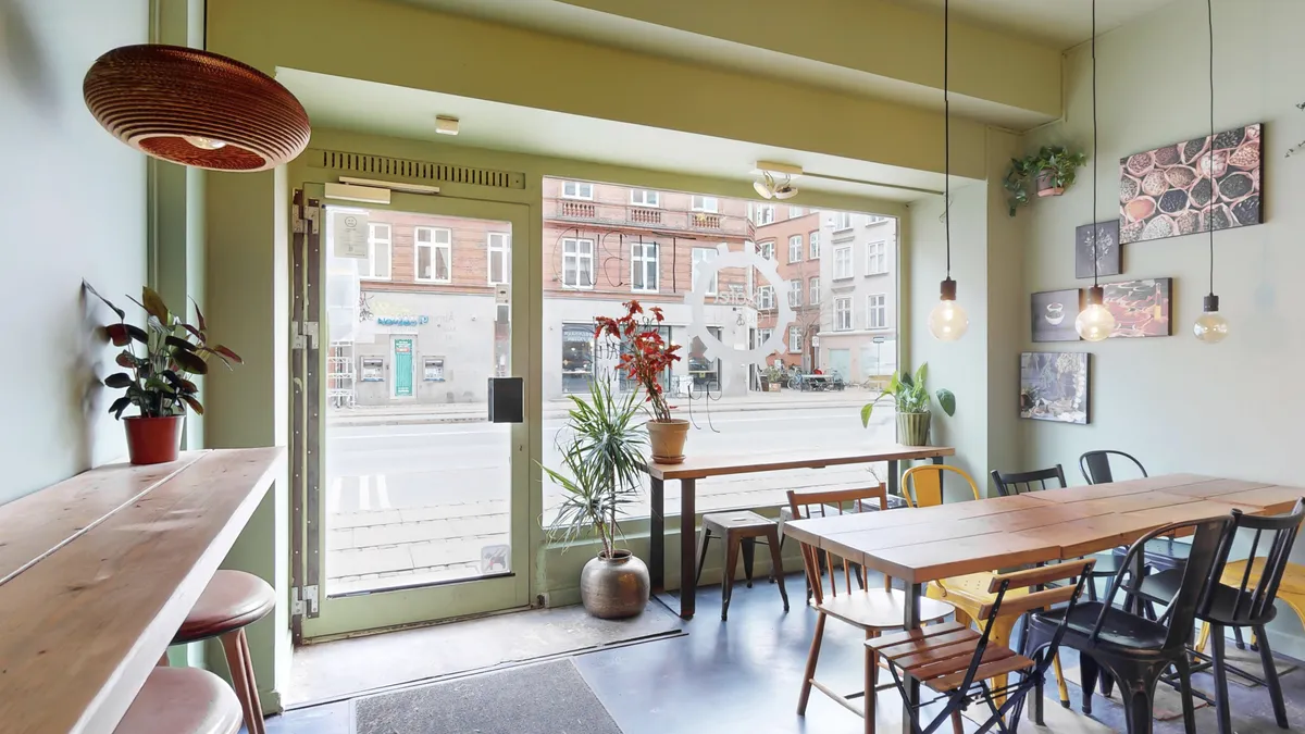En moderne restaurant med lyse, lyse farver, store vinduer og en indvendig plante. Bord og stole og en åben dør til gade