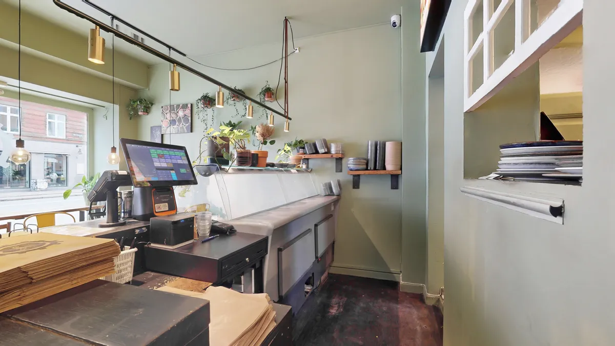 Køkken med POS system, computer og køkkenudstyr. Planter og hylder med glas og tallerkener. Vindue med udsigt til gaden.