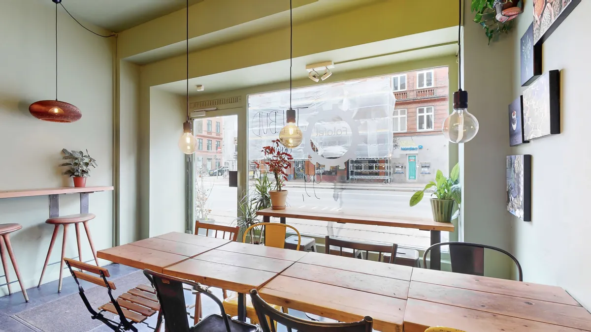 Store vinduer med skrift og tegninger samt lysekroner, borde og stole i restaurant med plantager og kunstværker på væggen