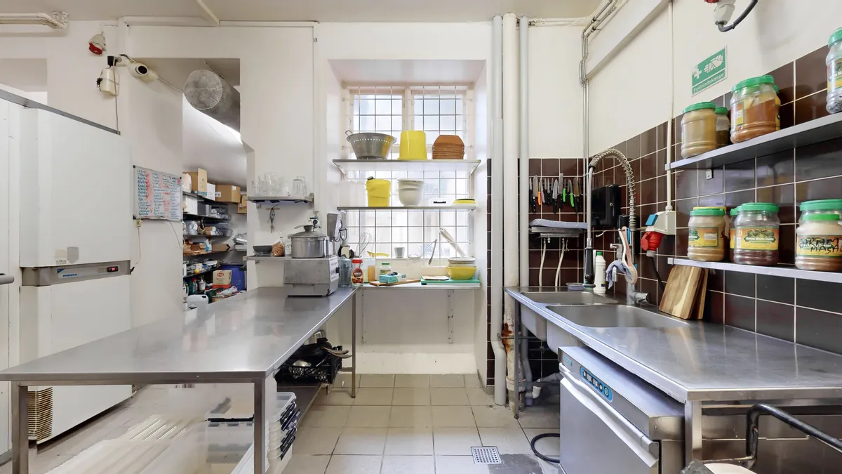 En ren og velorganiseret køkken med flere metalborde, en fryser, to vandhaner, to vaskebassiner og flere skabe og hylder med forskellige krydderier, sager og værktøj