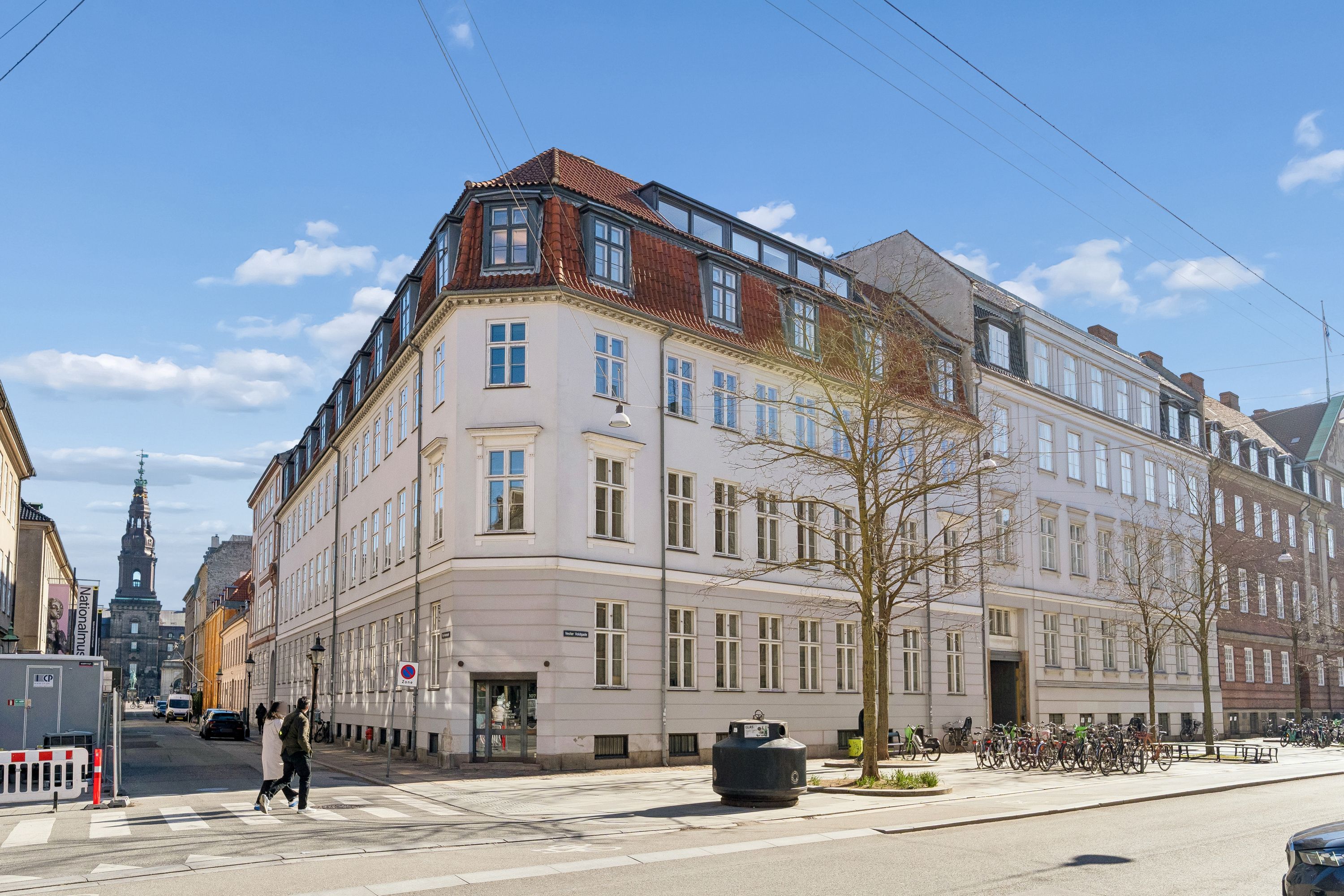 To personer krydser en gågade ved et hjørne af en bygning med mange vinduer og et rødt tag i København