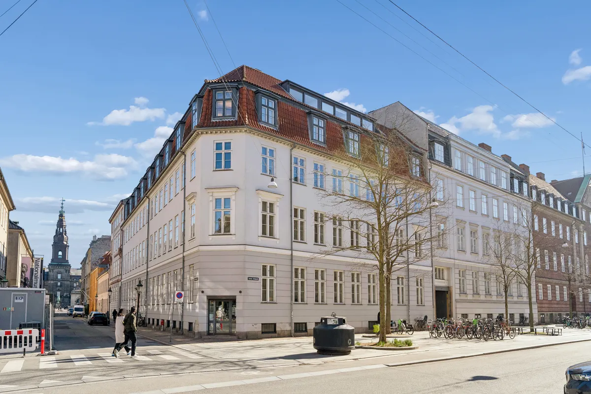 To personer krydser en gågade ved et hjørne af en bygning med mange vinduer og et rødt tag i København