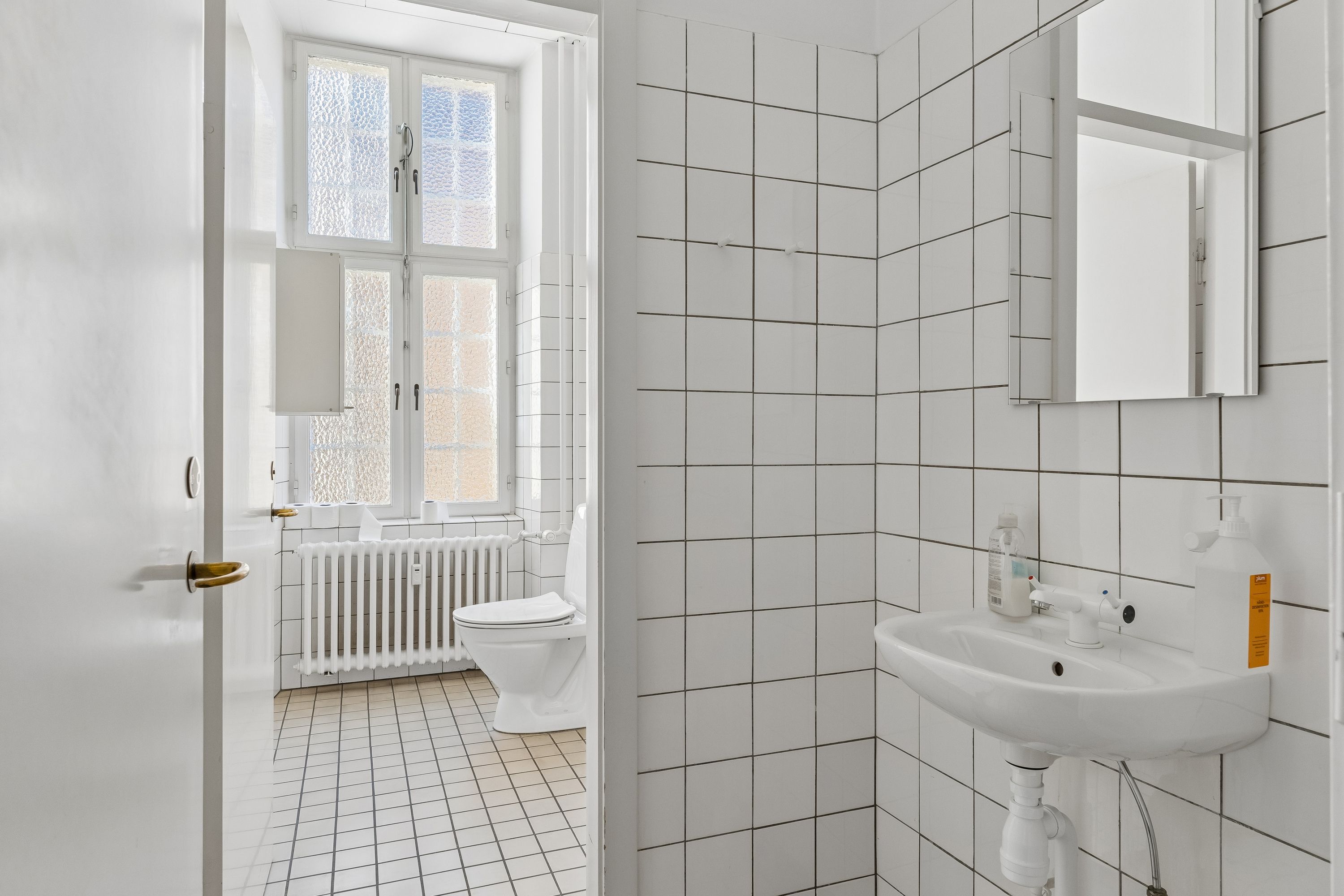Rent og rent hvidt badeværelse med toilet, radiator og håndvask med håndsæbe og desinfektionsmiddel