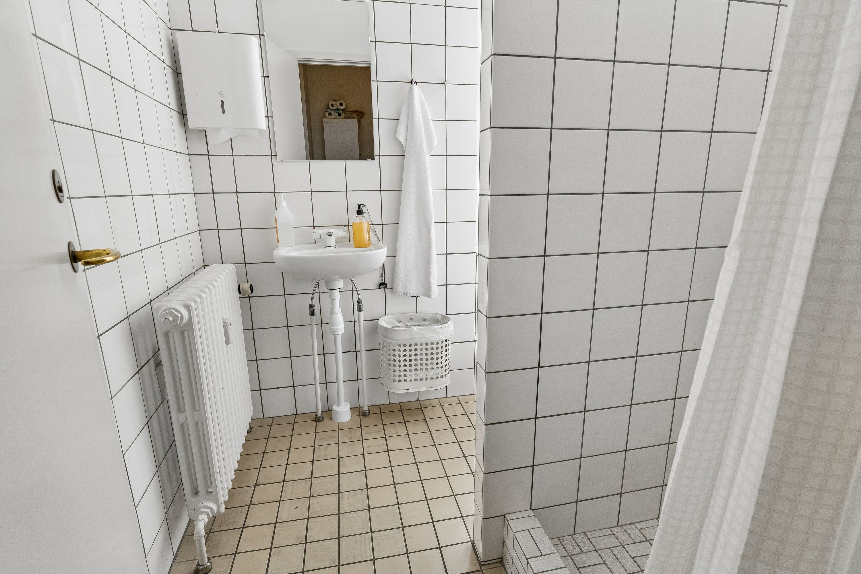 Kontor med hvidt toilet, hvidt badeværelse og brusekabine, radiator, hængeklæde og håndsæbe.