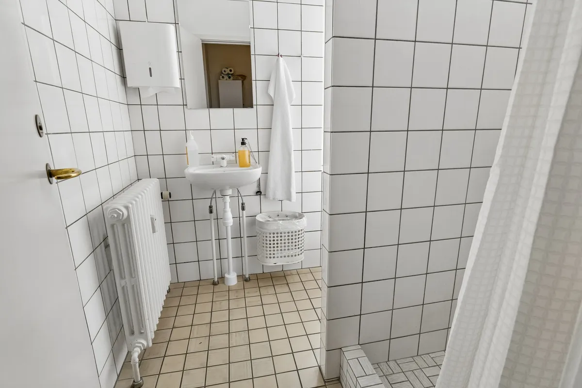 Kontor med hvidt toilet, hvidt badeværelse og brusekabine, radiator, hængeklæde og håndsæbe.