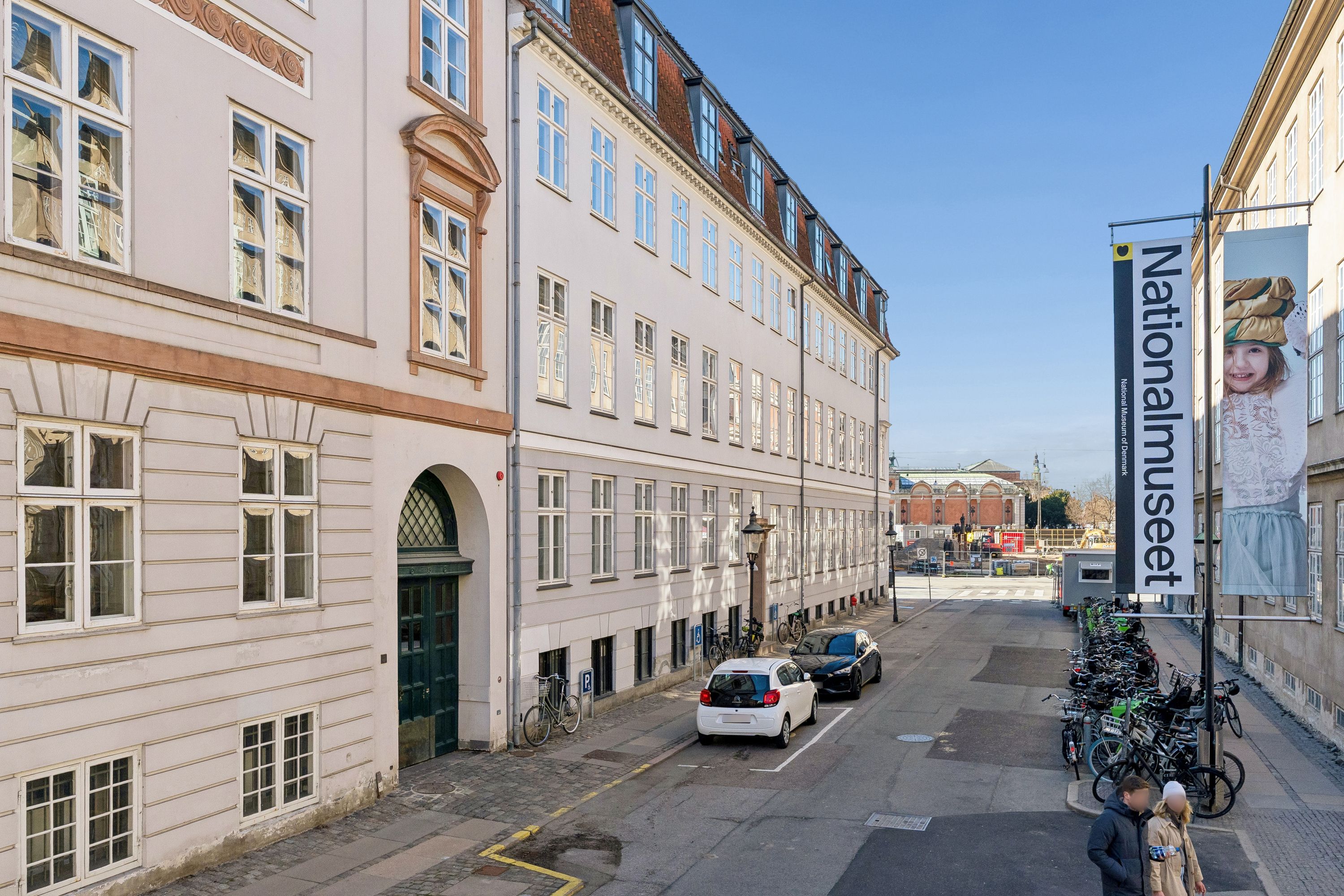 Kontor til leje i byen med glasruder og gamle bygninger. To biler parkeret og flere cykler står udenfor på gågaden. Et banner med teksten Nationalmuseet står på en stolpe