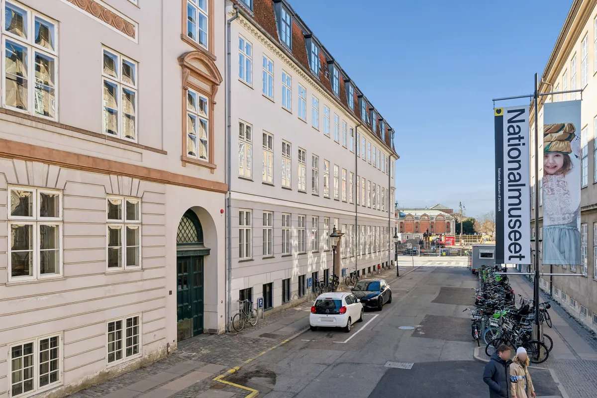 Kontor til leje i byen med glasruder og gamle bygninger. To biler parkeret og flere cykler står udenfor på gågaden. Et banner med teksten Nationalmuseet står på en stolpe
