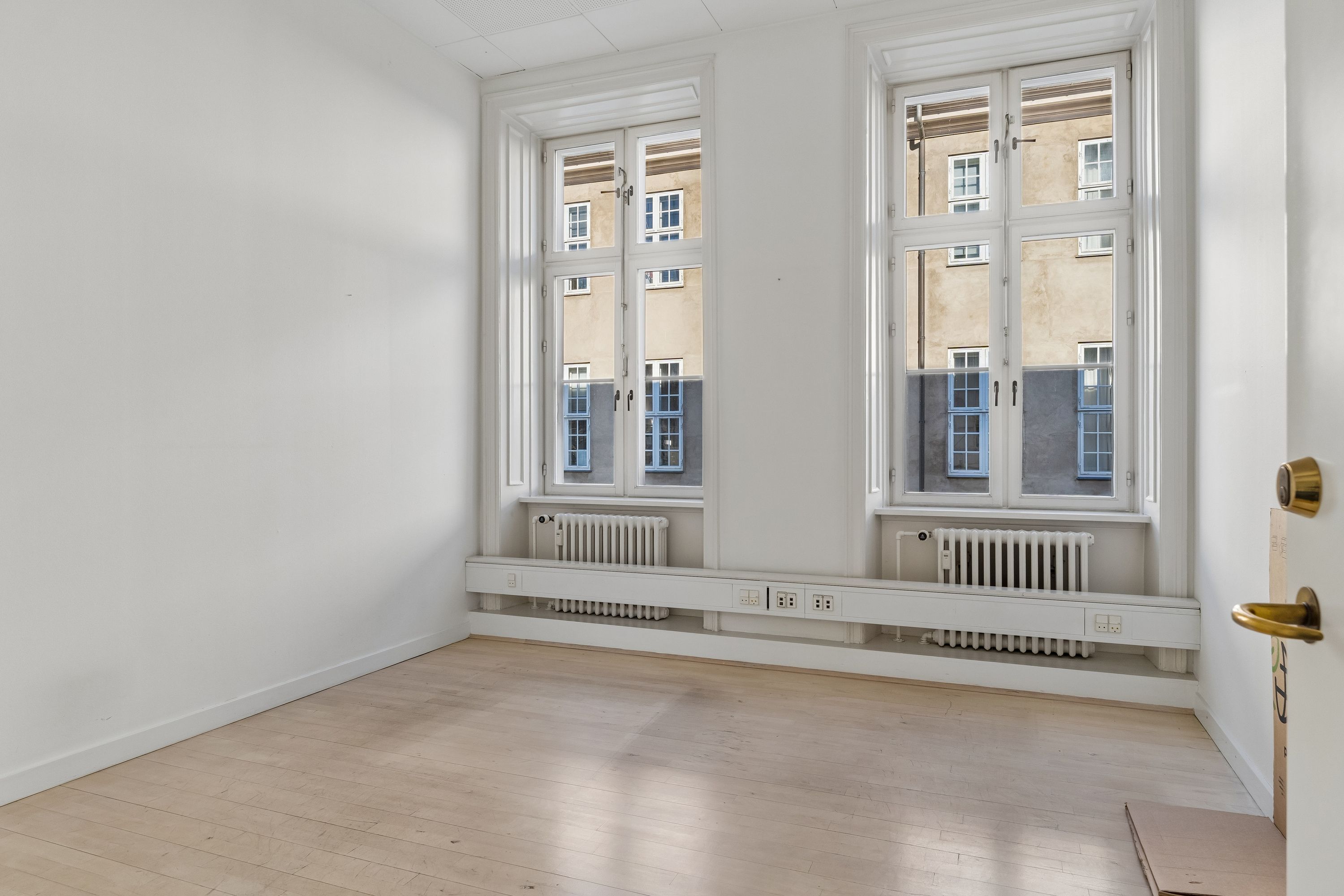 En tom kontorkontor med lysegul gulvbelægning, hvide vægge, to store vinduer og radiatorer under vinduet samt en lysegul dør med guldbeslag