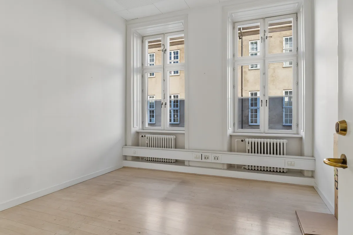 En tom kontorkontor med lysegul gulvbelægning, hvide vægge, to store vinduer og radiatorer under vinduet samt en lysegul dør med guldbeslag