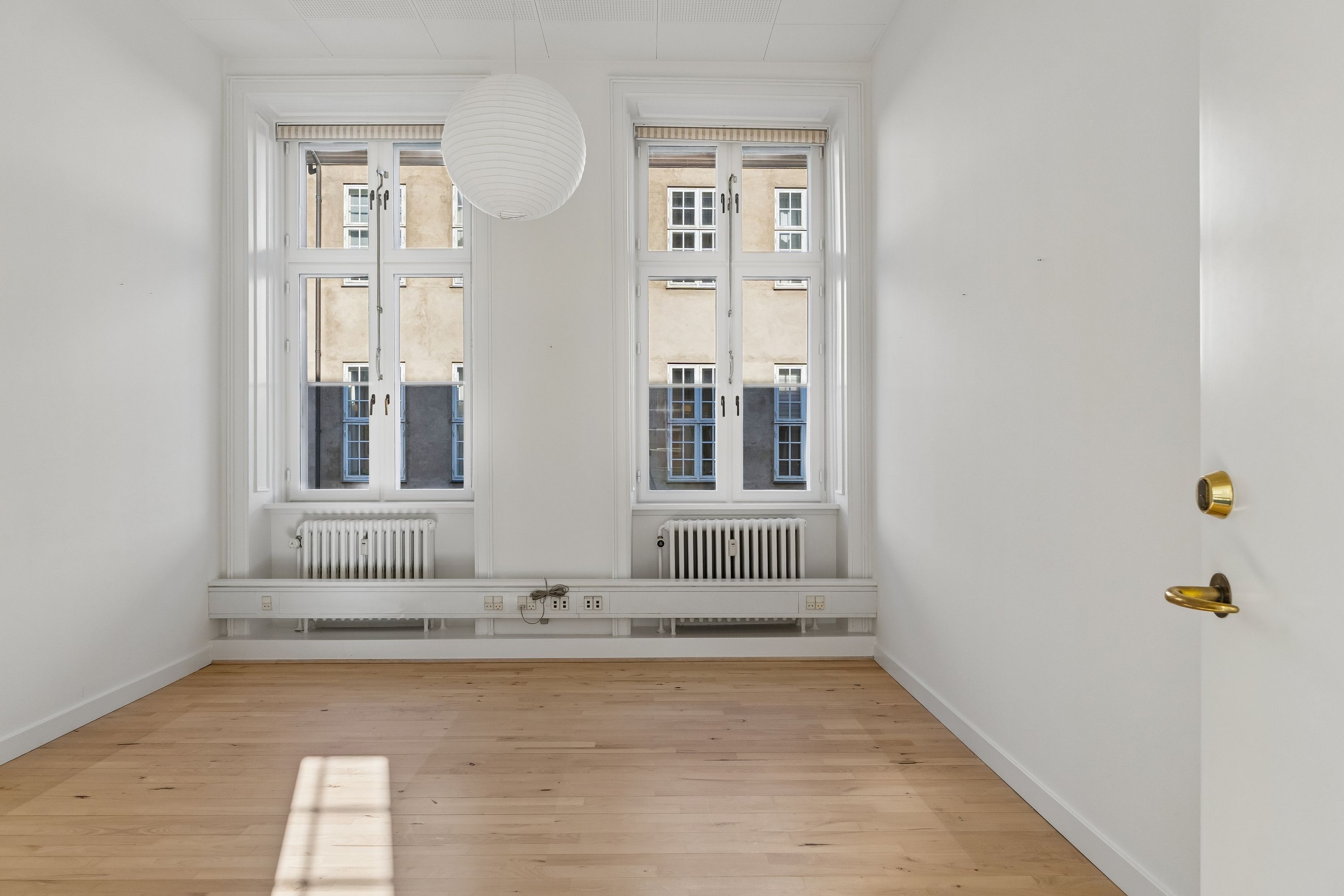 Kontor til leje med lyse gulve og hvide vægge, to store vinduer med gardiner, radiatorer og en hvid kuglelampe hængende fra loftet