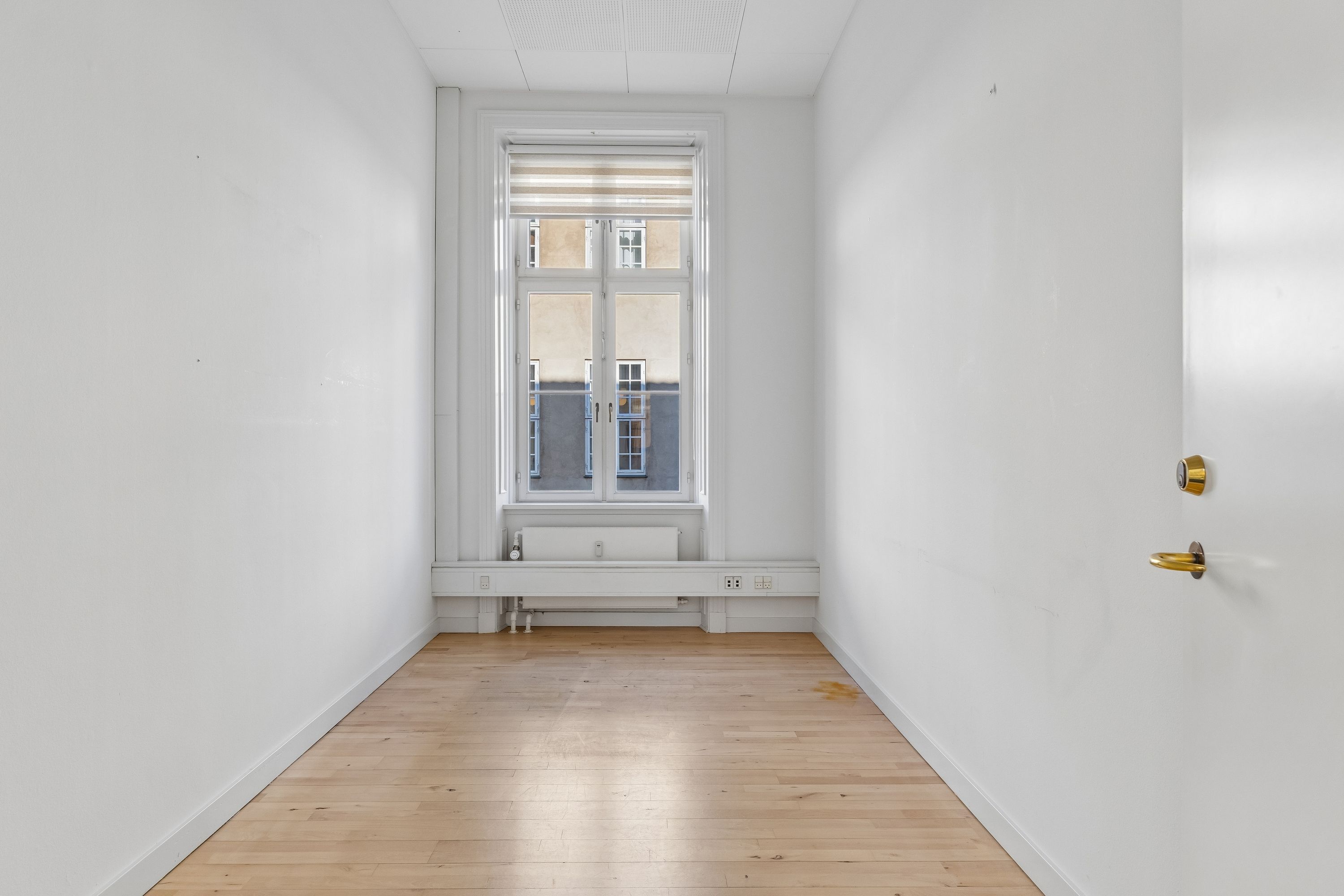 Et tomt kontorlokale med hvide vægge og lyse gulvplanker samt et vindue med en hvid gardin i en højbygning