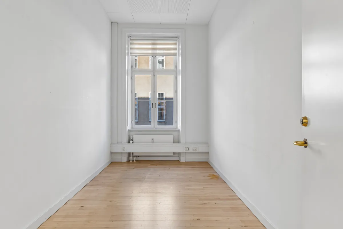 Et tomt kontorlokale med hvide vægge og lyse gulvplanker samt et vindue med en hvid gardin i en højbygning