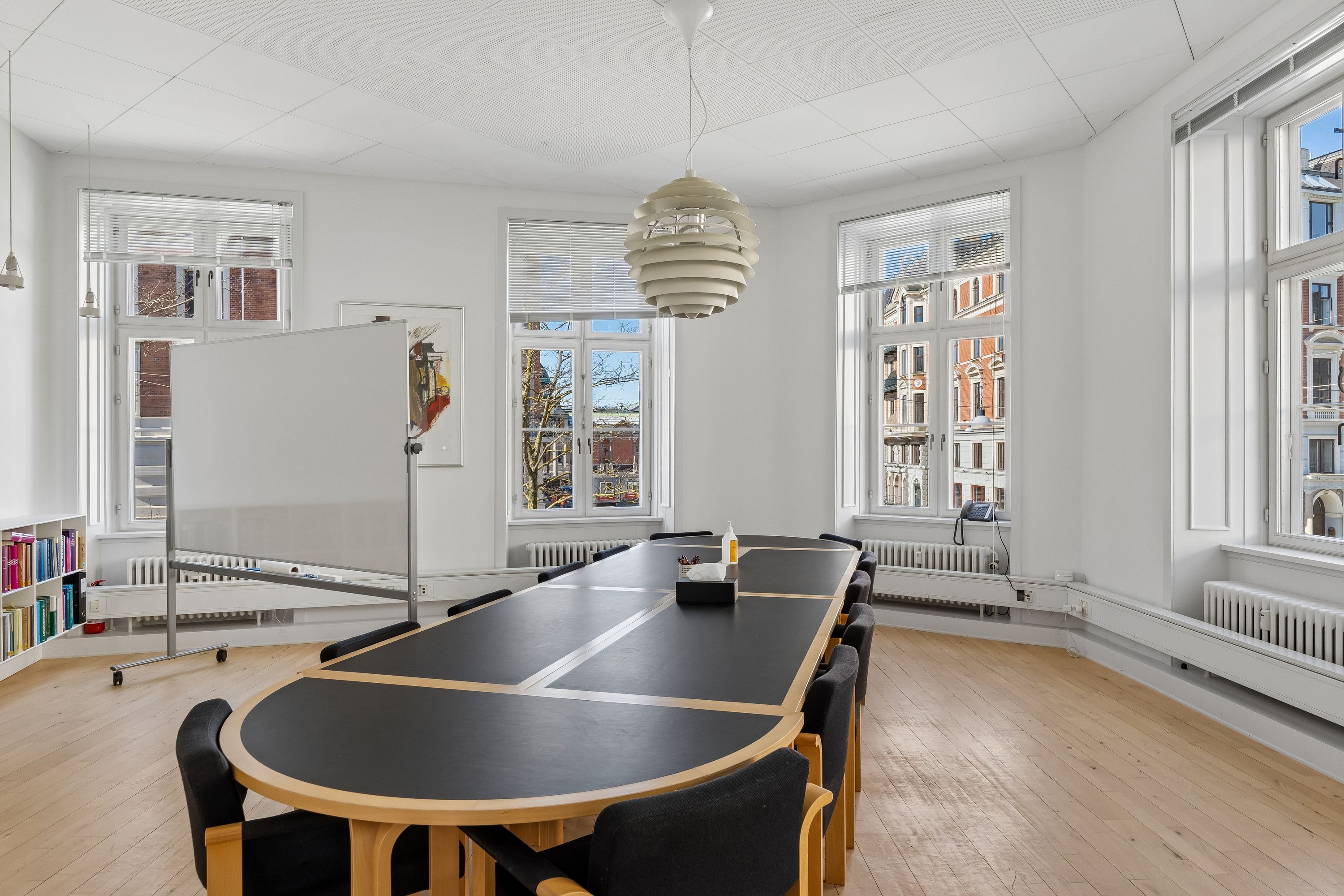 Kontor med lyse trægulve og møbler, to lange borde og en whiteboard samt lyse vinduer med gardiner