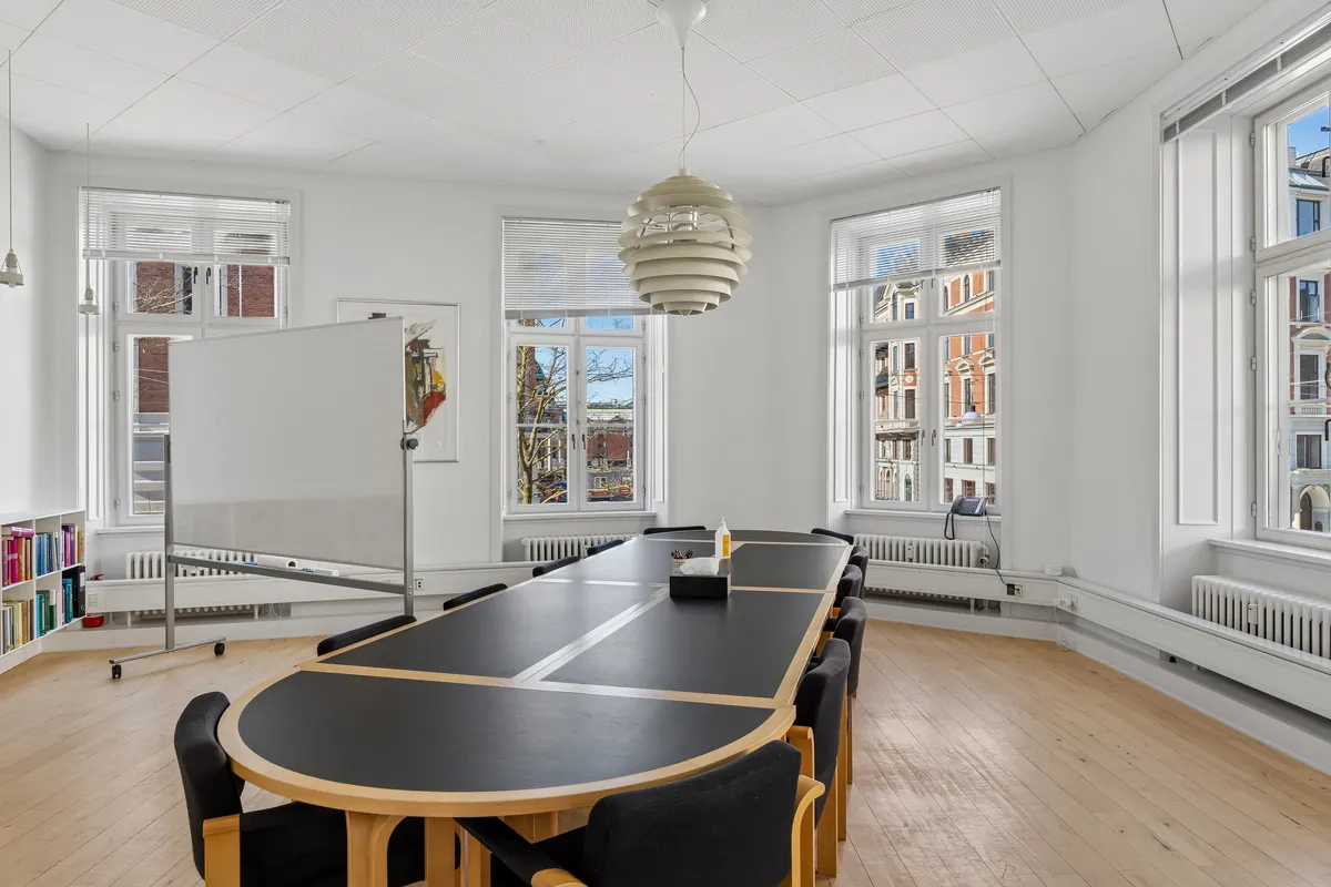 Kontor med lyse trægulve og møbler, to lange borde og en whiteboard samt lyse vinduer med gardiner